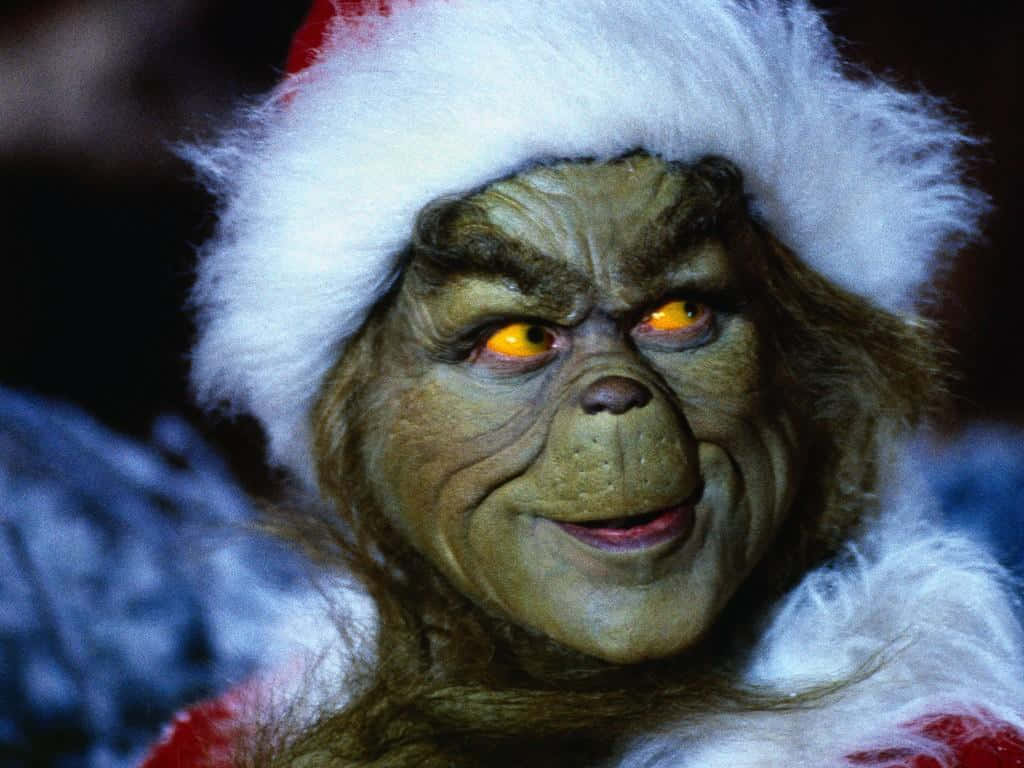 Christmas Grinch 桌布 壁紙 — 聖誕怪傑近照，戴著聖誕帽，發出橙色光芒的眼睛，帶著不高興的笑容。