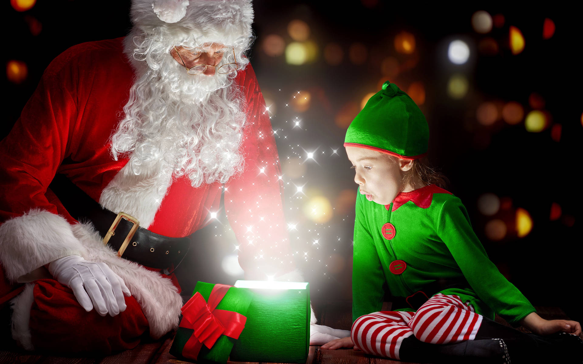 Santa Claus 桌布 壁紙 — 聖誕老人和一個小孩精靈好奇地凝視著一個發光的綠色禮物盒，在黑暗的散景背景下散發出閃爍的魔法。