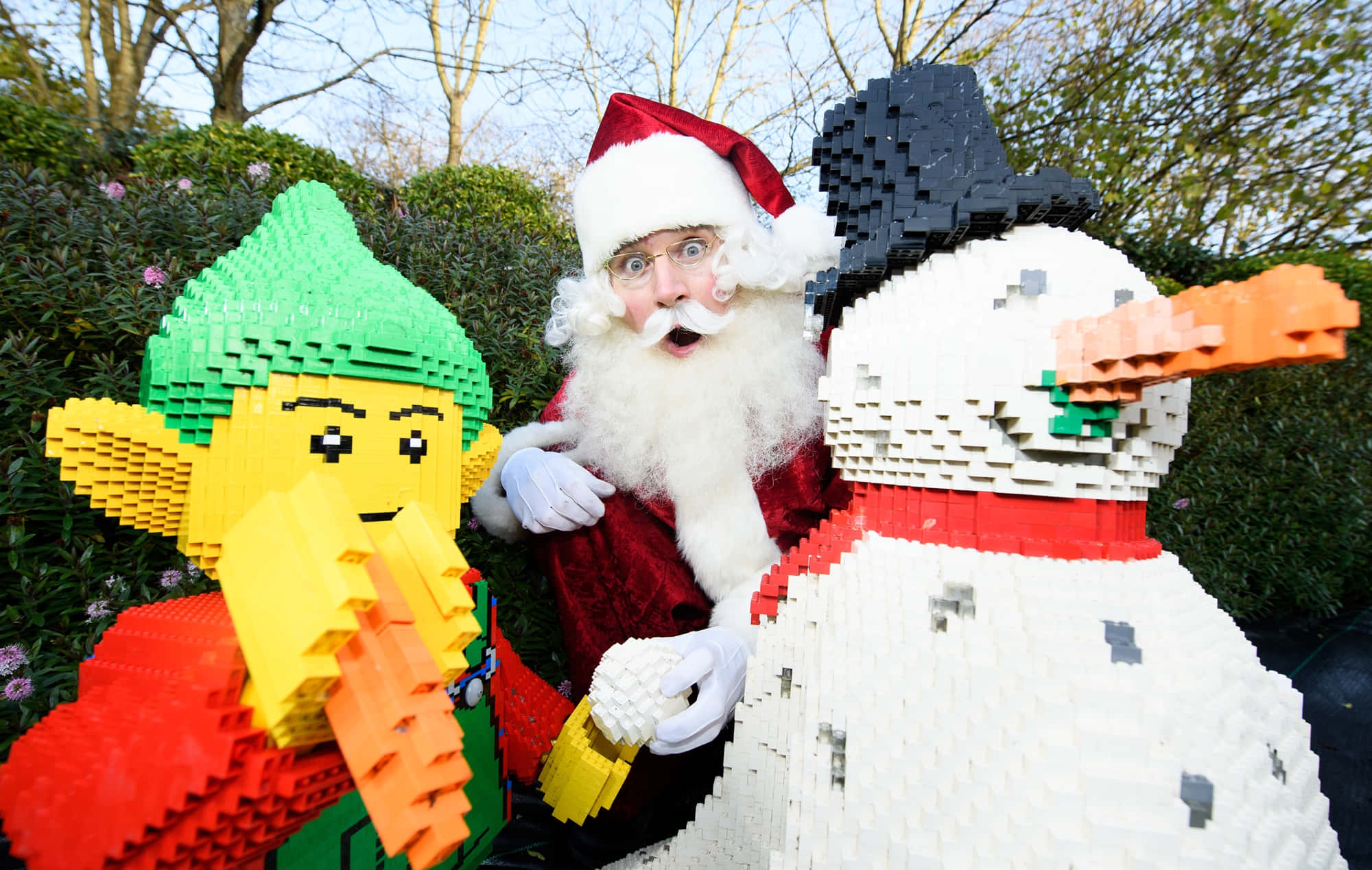 Legoland 桌布 壁紙 — 聖誕老人在一個充滿活力的戶外展示中，驚訝地從一個LEGO精靈和雪人之間探出頭來。