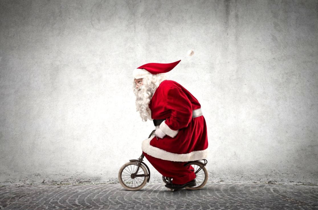 Santa Claus 桌布 壁紙 — 聖誕老人在鵝卵石街道上，在有紋理的灰色牆前騎著一輛小自行車。