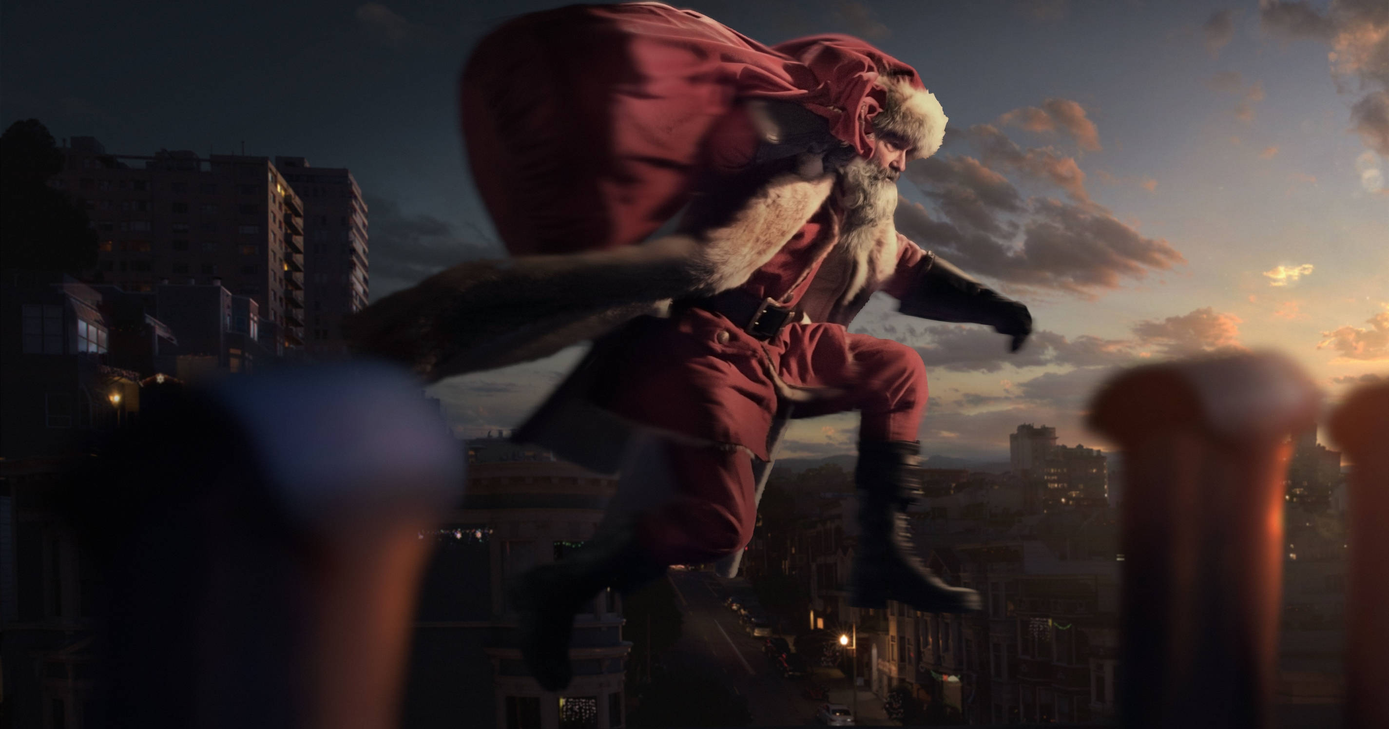 Christmas Movie 桌布 壁紙 — 聖誕老人在黃昏時分翱翔於城市天際之上，背著一個巨大的禮物袋，映襯著溫暖的橙色和紫色天空。
