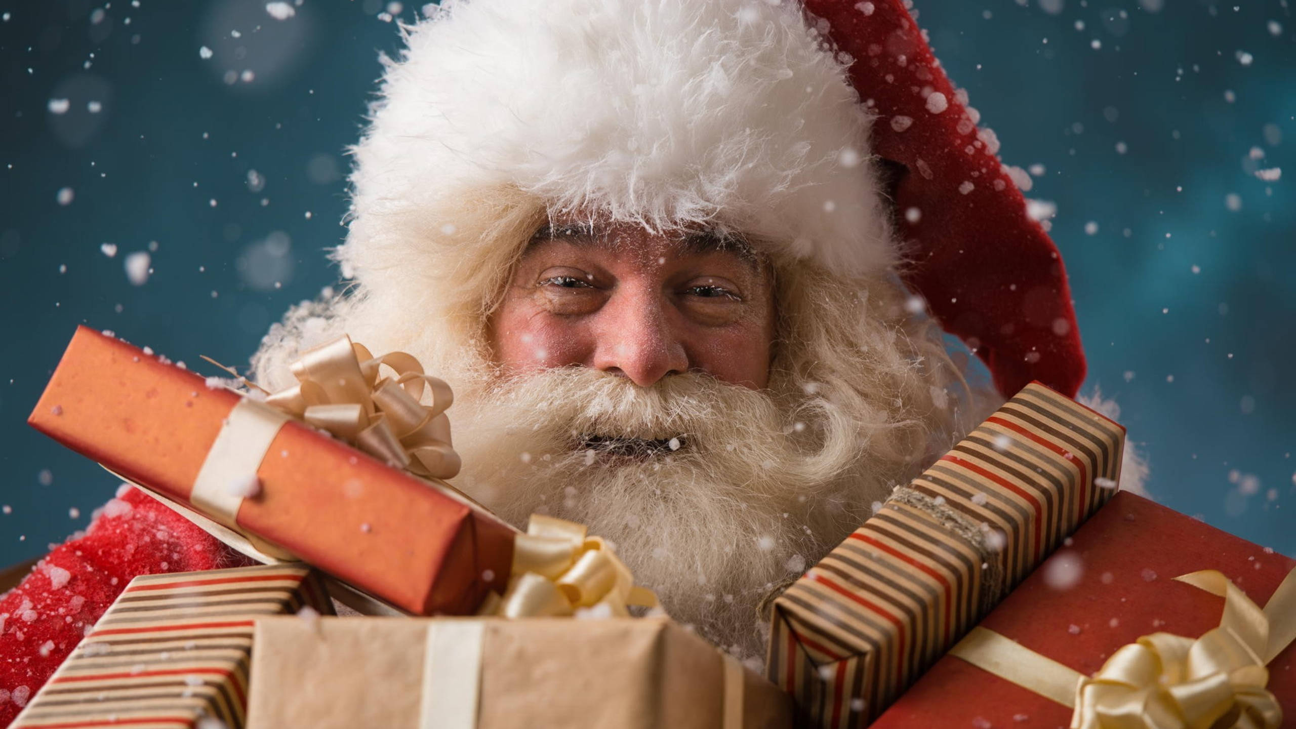 Santa Claus 桌布 壁紙 — 聖誕老人帶著溫暖的笑容從一堆包裝好的禮物後面探出頭來，雪花在深藍色背景上飄落。
