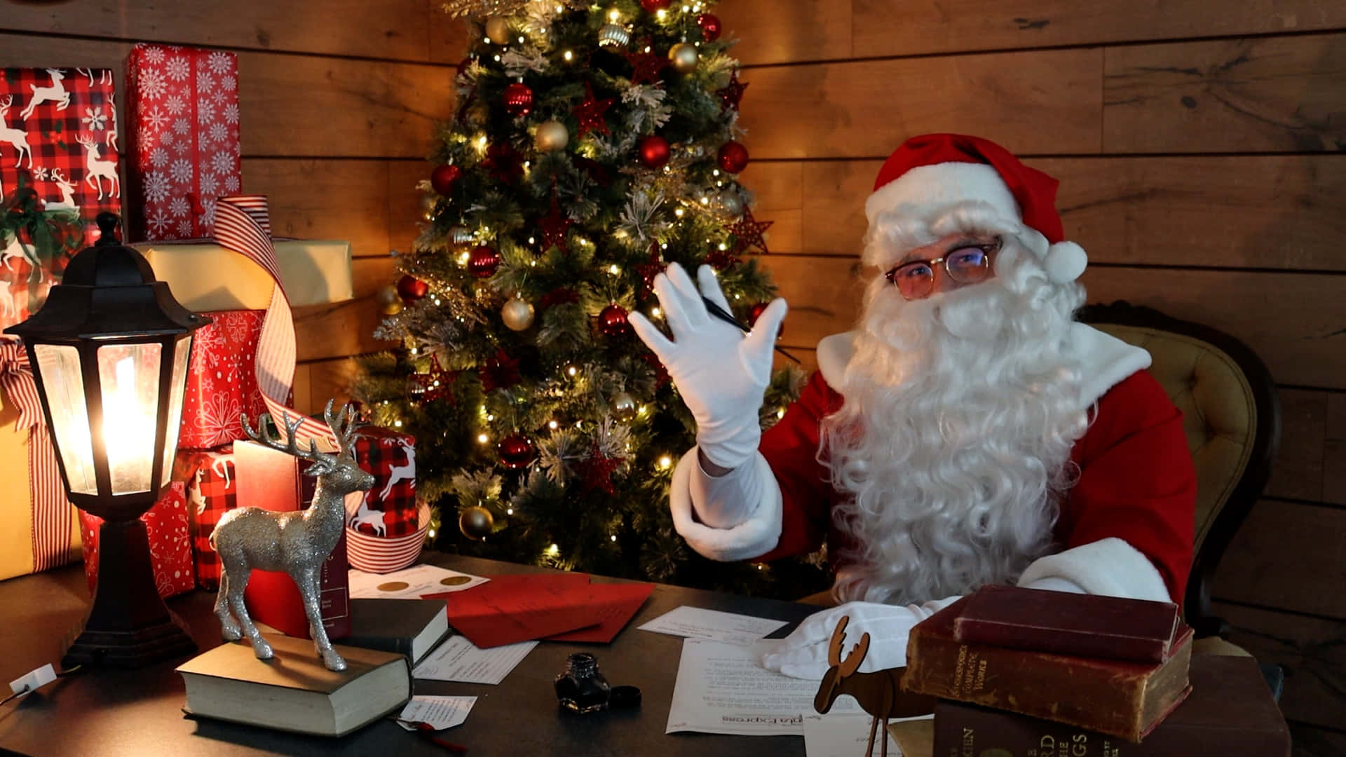 Christmas Virtual 桌布 壁紙 — 聖誕老人從他的木桌旁愉快地揮手，旁邊是一棵閃爍的聖誕樹，裝飾著紅色飾物和閃爍的燈光。
