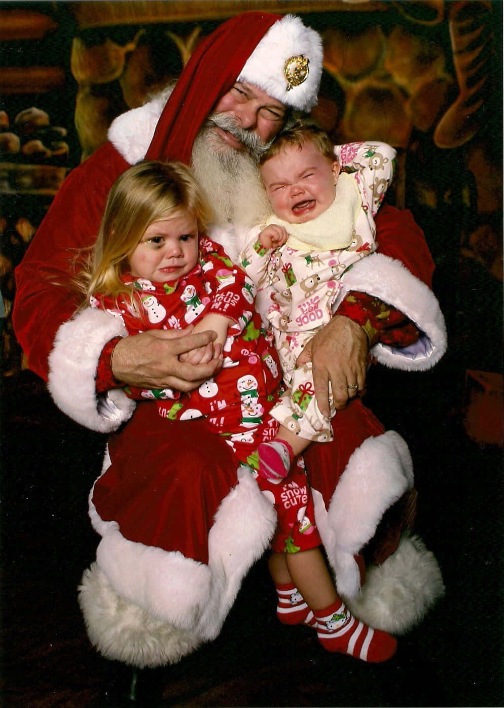 Santa Claus 桌布 壁紙 — 聖誕老人抱著一個哭泣的嬰兒和一個看起來悲傷的女孩，兩人穿著節日睡衣，背景是深色木板。