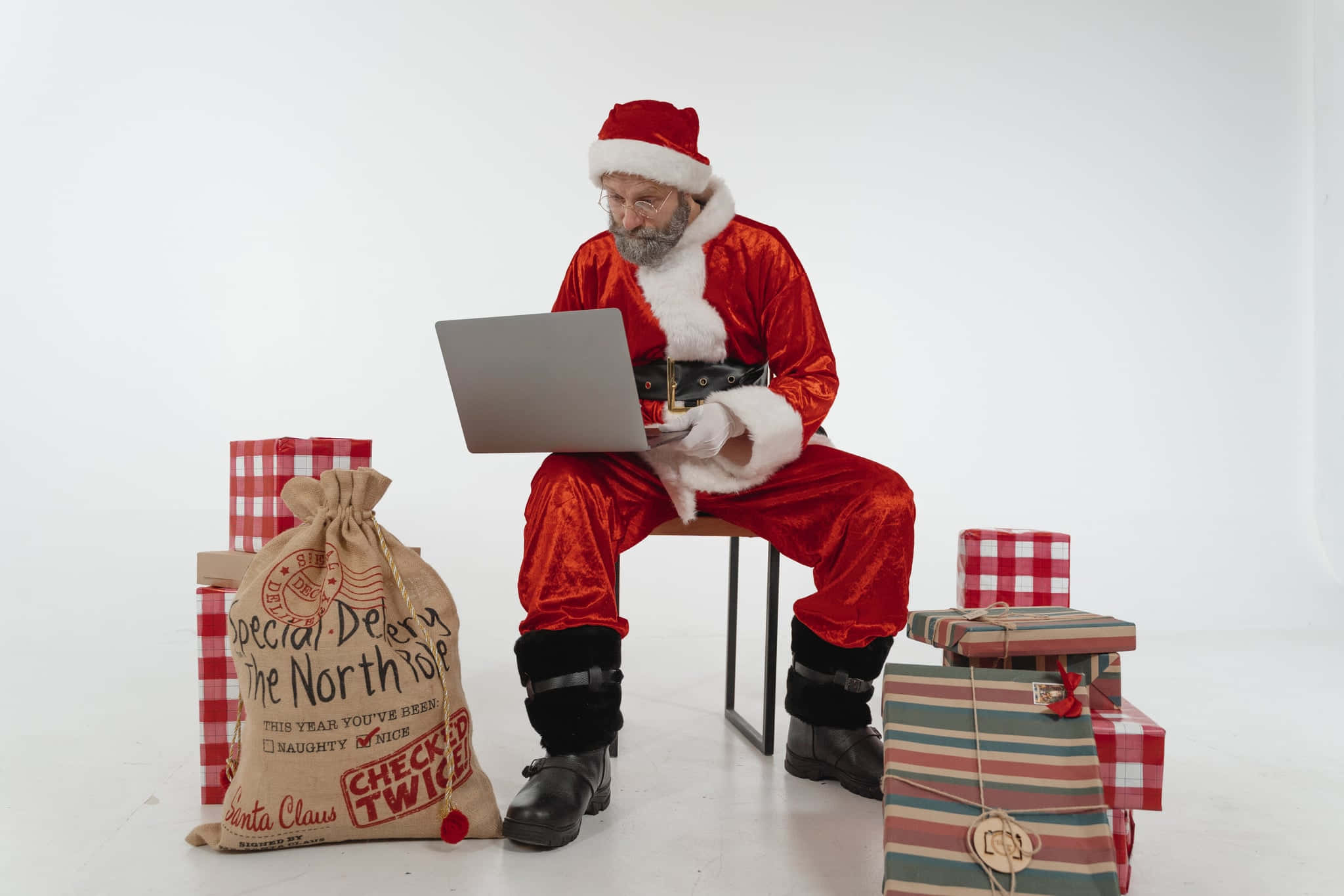 Santa Claus 桌布 壁紙 — 聖誕老人穿著紅色天鵝絨套裝坐在凳子上使用筆記型電腦，周圍是包裝好的禮物和一個標有「北極特別送遞」的袋子。