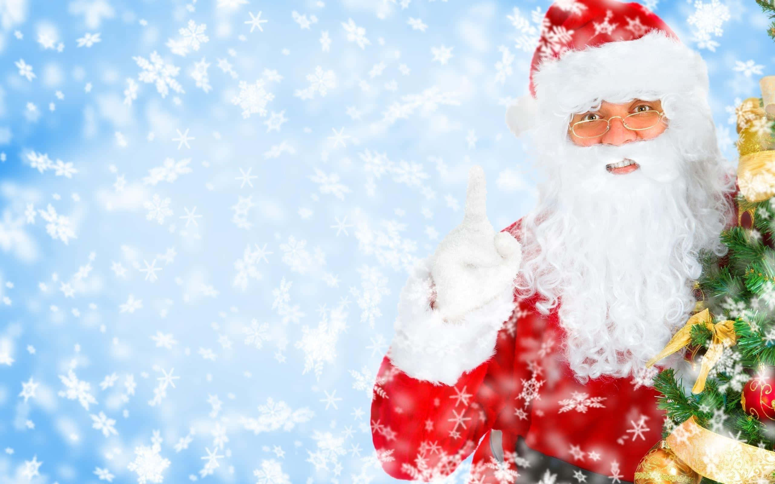Santa Claus 桌布 壁紙 — 聖誕老人穿著紅色西裝，在裝飾好的樹旁愉快地指著，雪花在明亮的藍天下飄落。