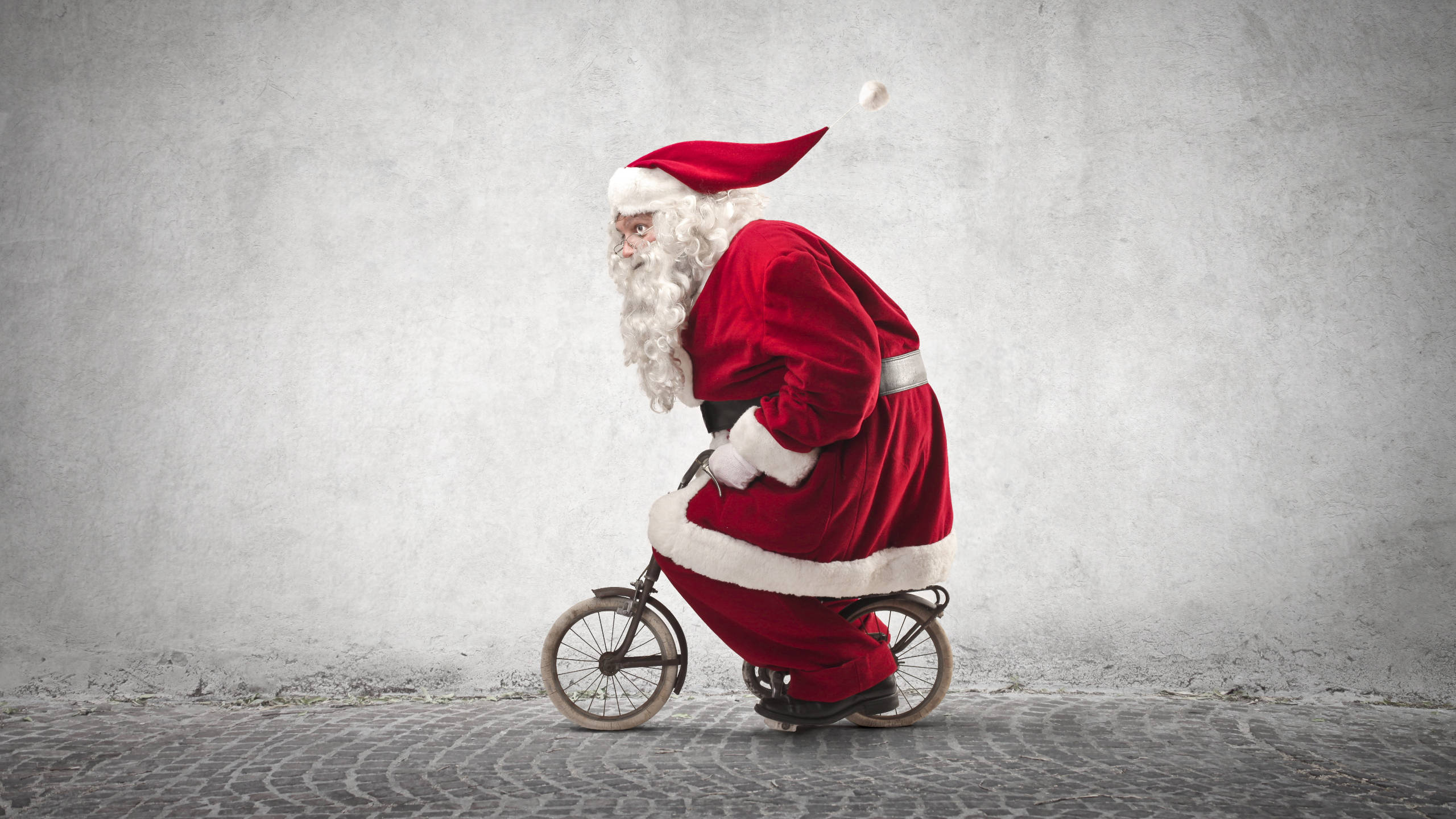 Santa Claus 桌布 壁紙 — 聖誕老人穿著紅色西裝，在鵝卵石街道上對著有紋理的灰色牆壁騎著一輛小型自行車。
