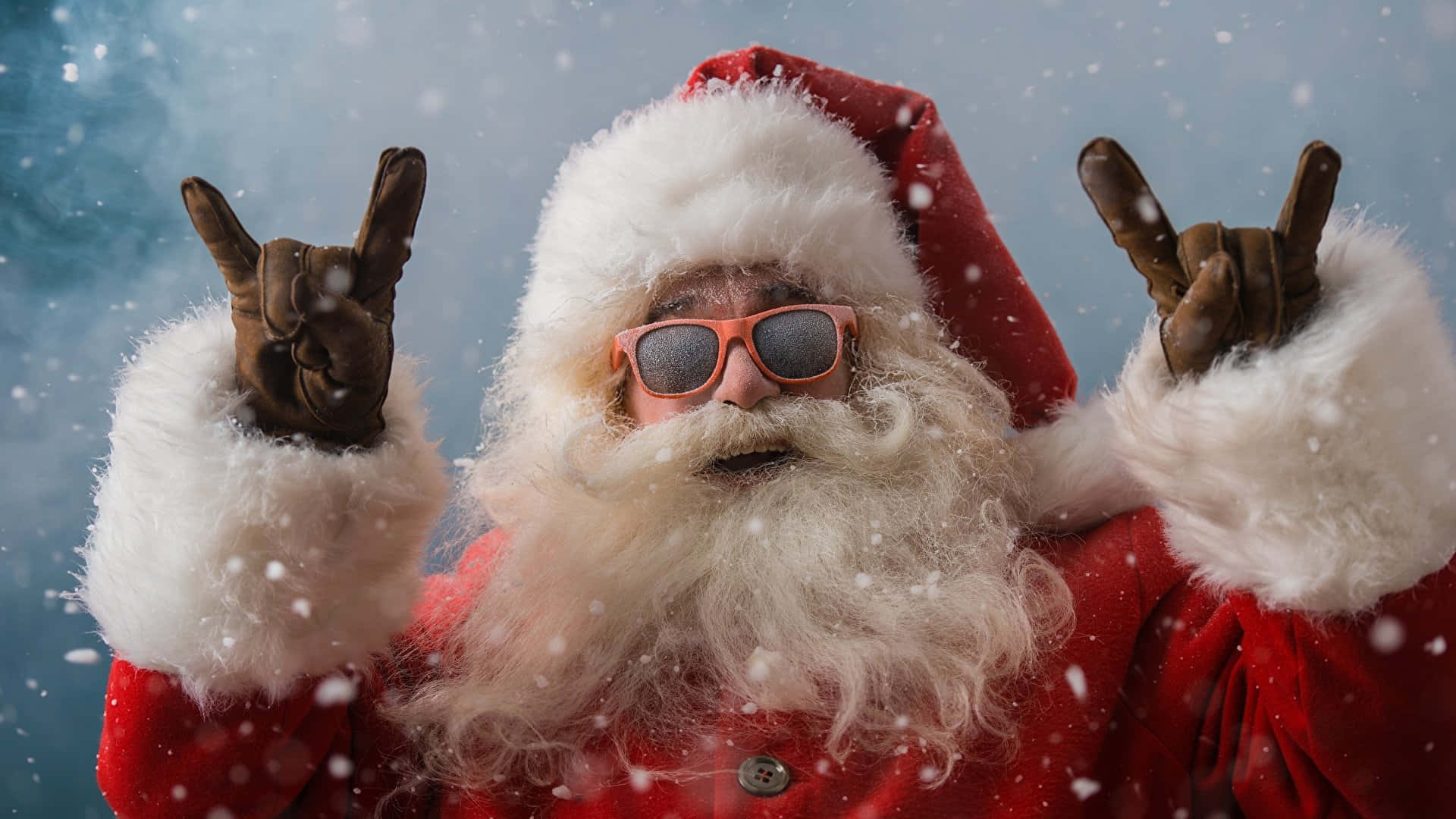 Santa Claus 桌布 壁紙 — 聖誕老人穿著紅色西裝和太陽眼鏡，在藍天背景下於飄雪中做出搖滾手勢。