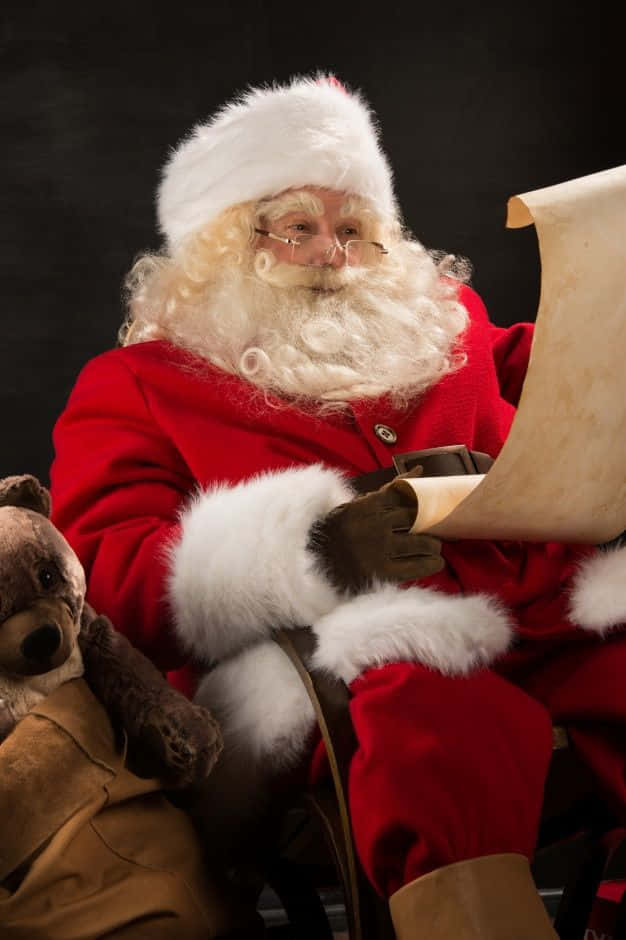 Santa Claus 桌布 壁紙 — 聖誕老人身穿紅色白邊套裝，坐在深色背景前的棕色泰迪熊旁邊閱讀卷軸。