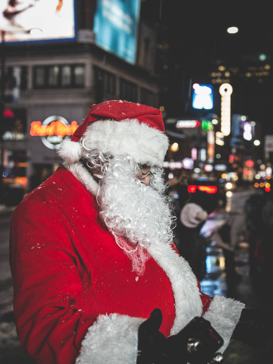 Santa Claus 桌布 壁紙 — 聖誕老人身穿紅色西裝，帽子上有雪花，站在夜晚繁華的城市中，身後是發光的廣告牌。