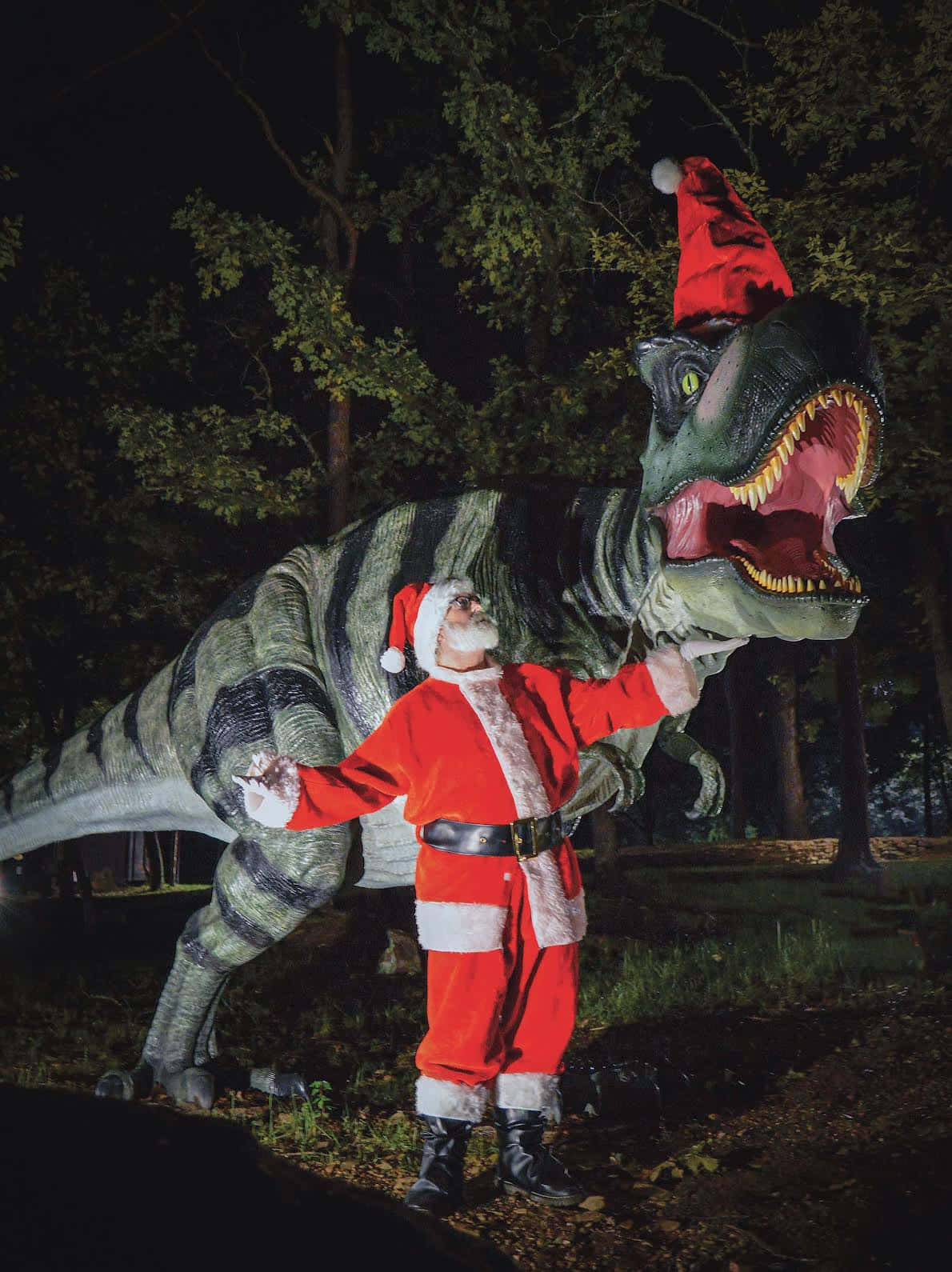 Dino 桌布 壁紙 — 聖誕老人身穿紅色西裝和帽子，站在夜晚黑暗森林中咆哮的恐龍模型旁邊。