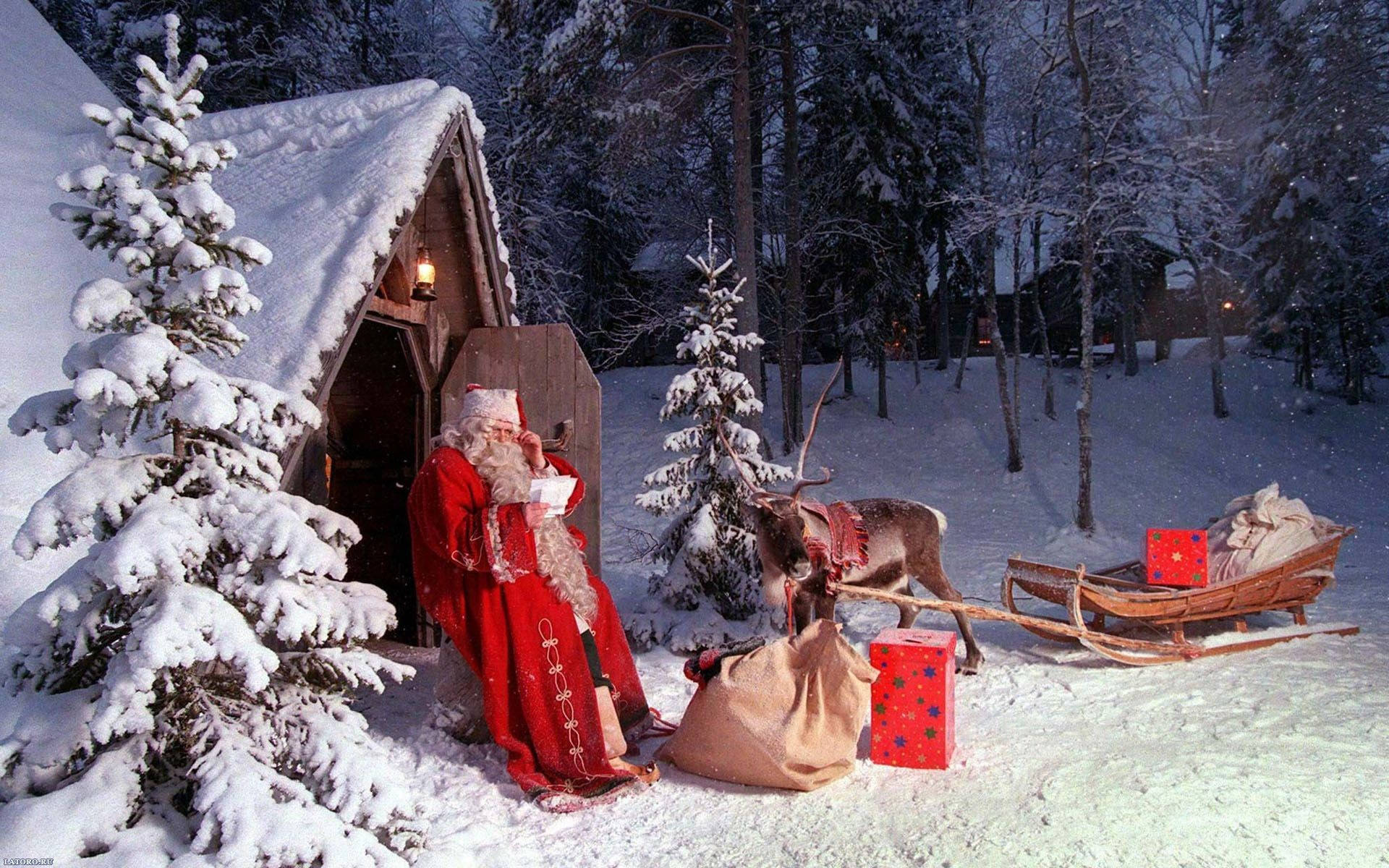 christmas holiday desktop 桌布 壁紙 — 聖誕老人身穿紅色長袍，在黃昏的冬季森林中，站在一間被雪覆蓋的小屋外，旁邊有一隻馴鹿和雪橇。