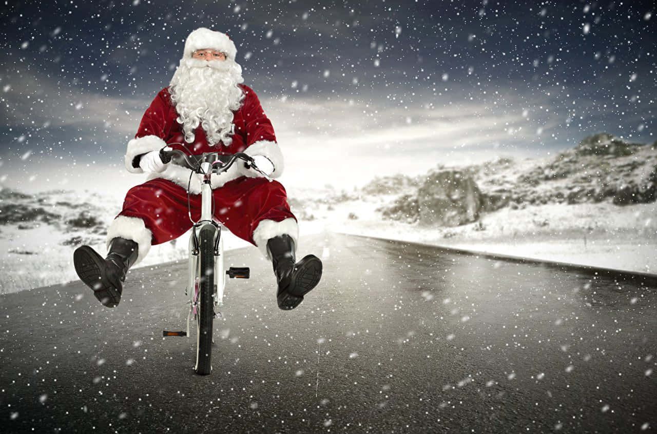 聖誕節 桌布 壁紙 — 聖誕老人騎著腳踏車行駛在積雪的道路上，雪花飄落，背景是白雪覆蓋的山丘。