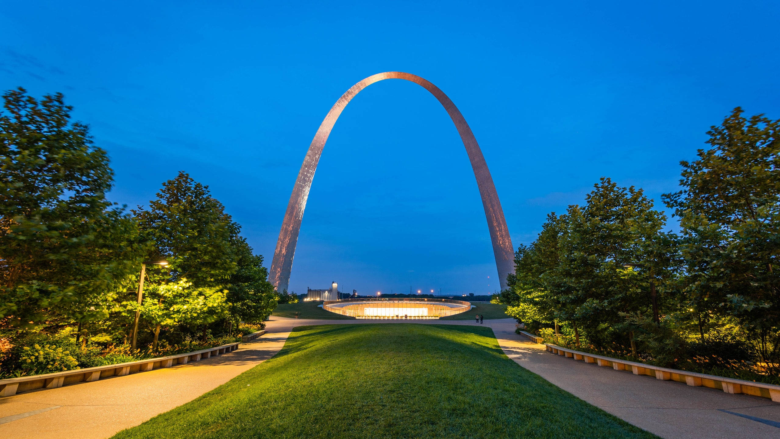 Architecture 桌布 壁紙 — 聖路易斯拱門在深藍色暮色天空下溫暖地發光，周圍環繞著茂密的綠樹和蜿蜒的小徑。