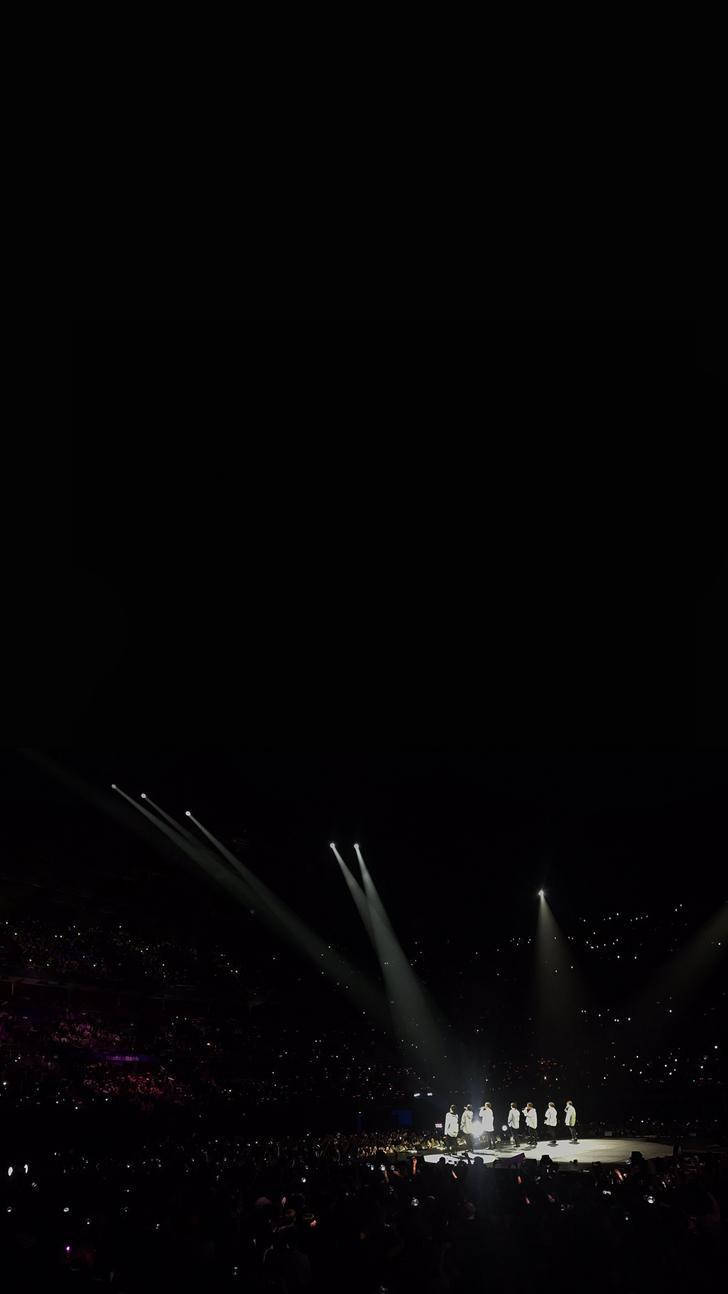 Bts Black 桌布 壁紙 — 聚光燈光束劃破黑暗，照射在音樂會舞台上表演者與觀眾舉起發光手機的海洋上。