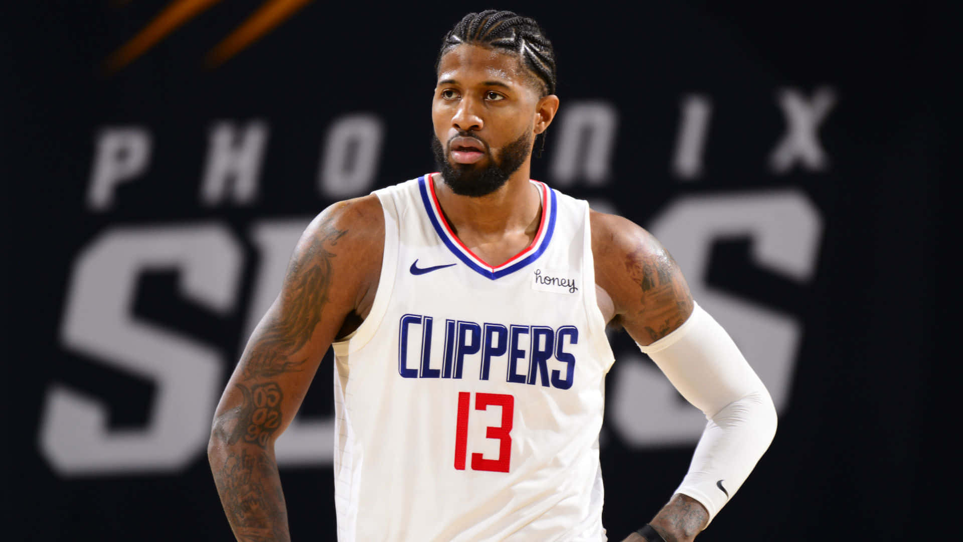 Paul George Clippers 桌布 壁紙 — 職業籃球運動員保羅·喬治身穿白色快艇隊球衣（13號），在帶有模糊球隊品牌的深色背景下自信地站立。
