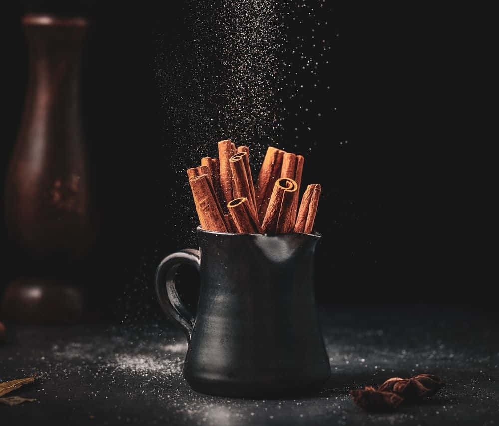 shoot 桌布 壁紙 — 肉桂棒和粉末從黑色陶瓷杯傾瀉而出，背景是深色背景，後面有一個模糊的木花瓶。
