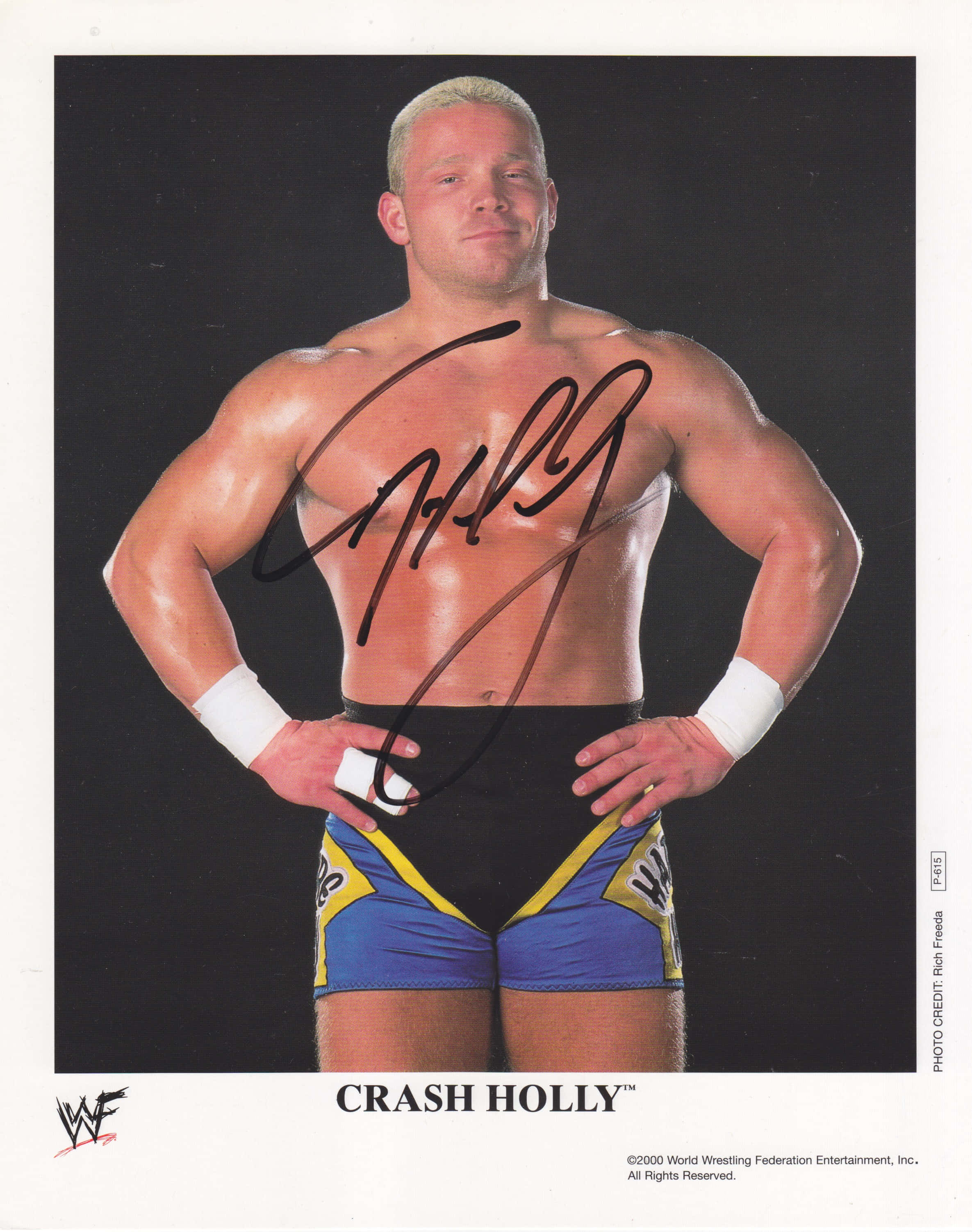 Crash Holly 桌布 壁紙 — 肌肉發達的摔角手 Crash Holly 自信地雙手叉腰站在黑色背景前，穿著藍黃色短褲和白色護腕。