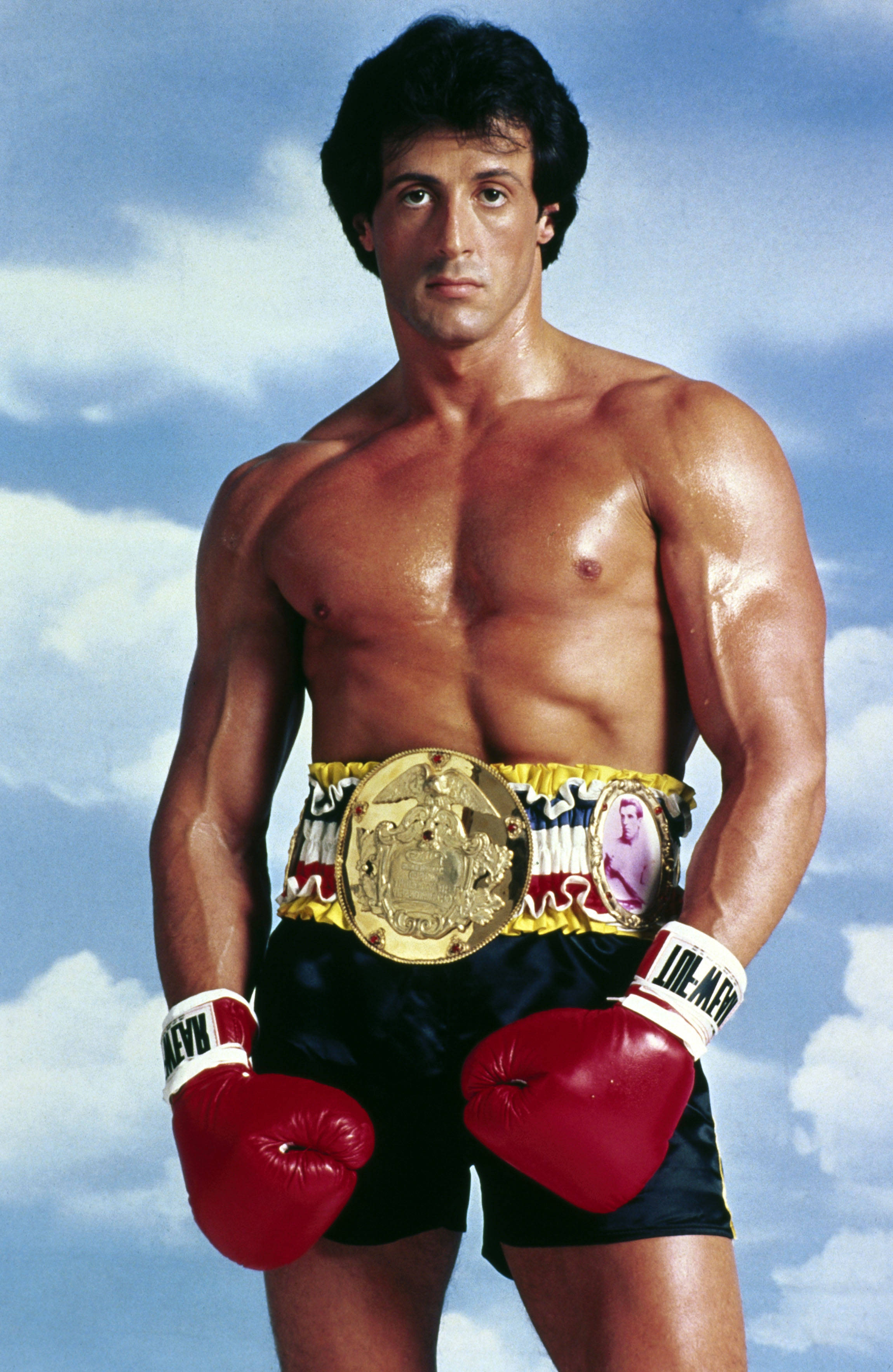 rocky 桌布 壁紙 — 肌肉發達的男人戴著紅色拳擊手套和冠軍腰帶，在藍天白雲下自信地站立。