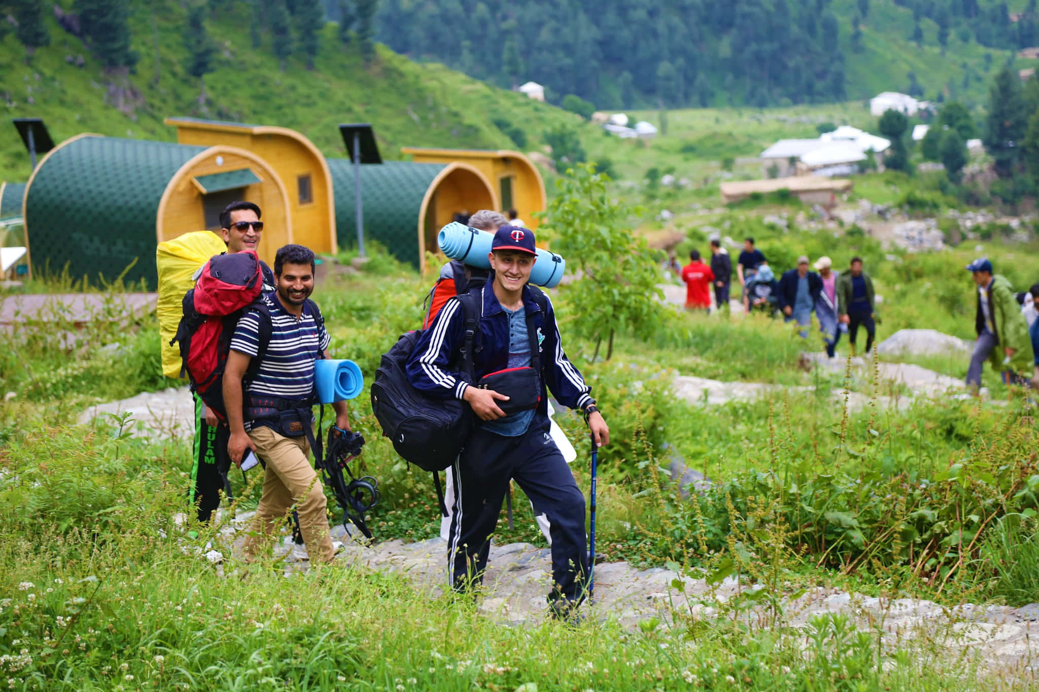 Eco-tourism 桌布 壁紙 — 背著背包和登山杖的遠足者，走過山谷中色彩繽紛的圓頂小屋附近茂密的綠草。