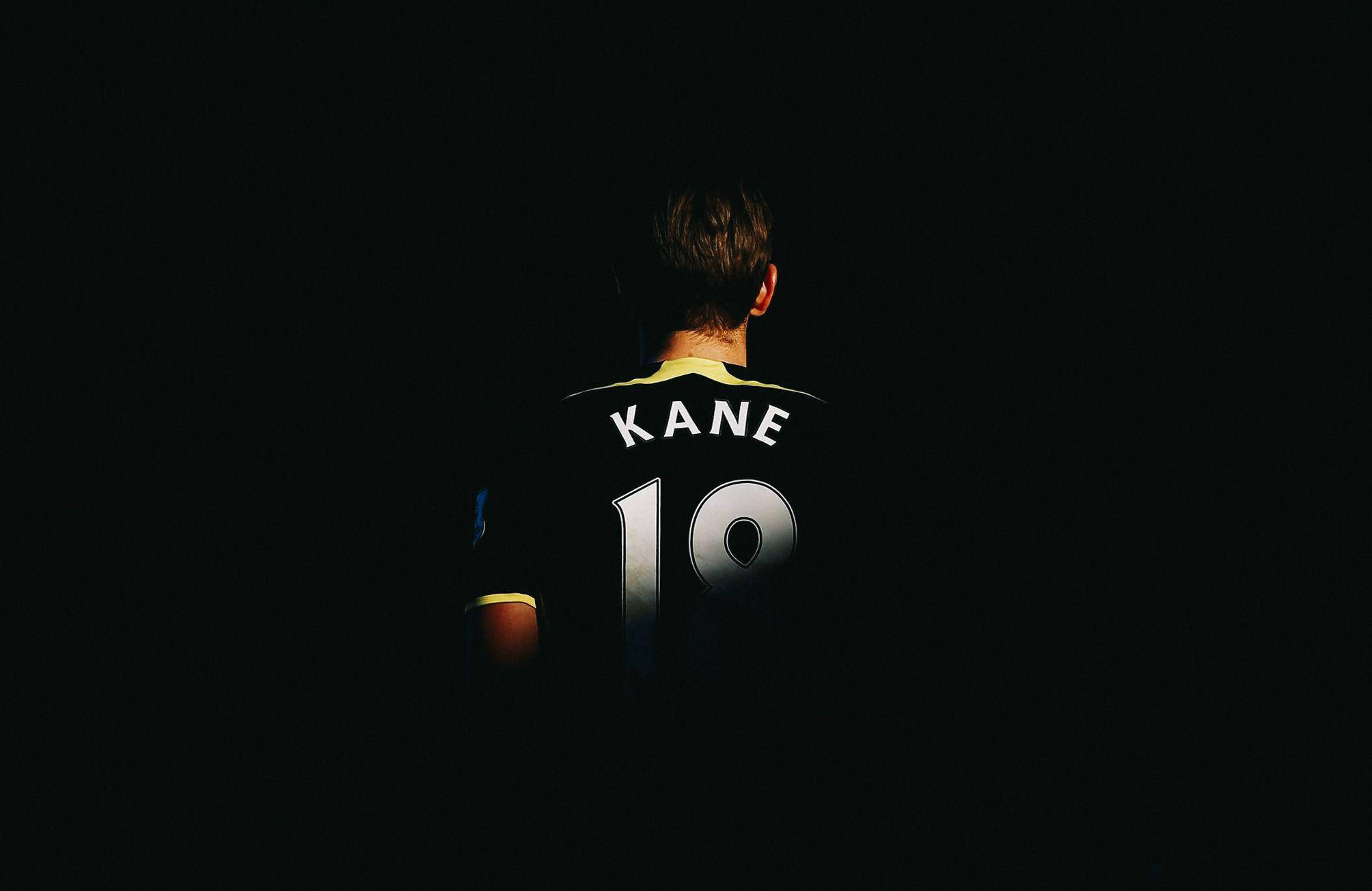 哈里·凱恩 桌布 壁紙 — 背面視圖，一名足球員穿著黑色 'KANE 19' 球衣，背景黑暗，燈光戲劇性。