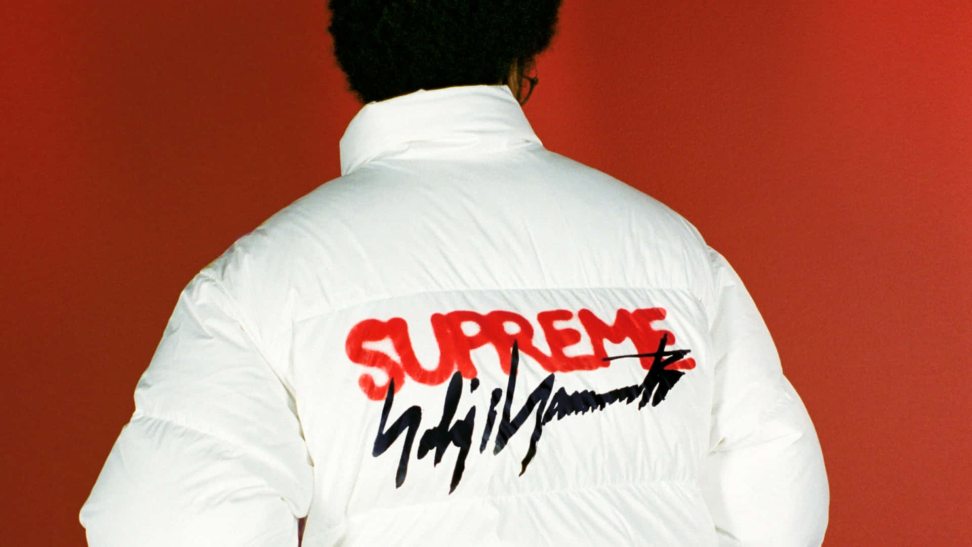 supreme 桌布 壁紙 — 背面視角，一個人穿著白色羽絨外套，上有紅色Supreme標誌和黑色簽名，背景為純紅色牆壁。