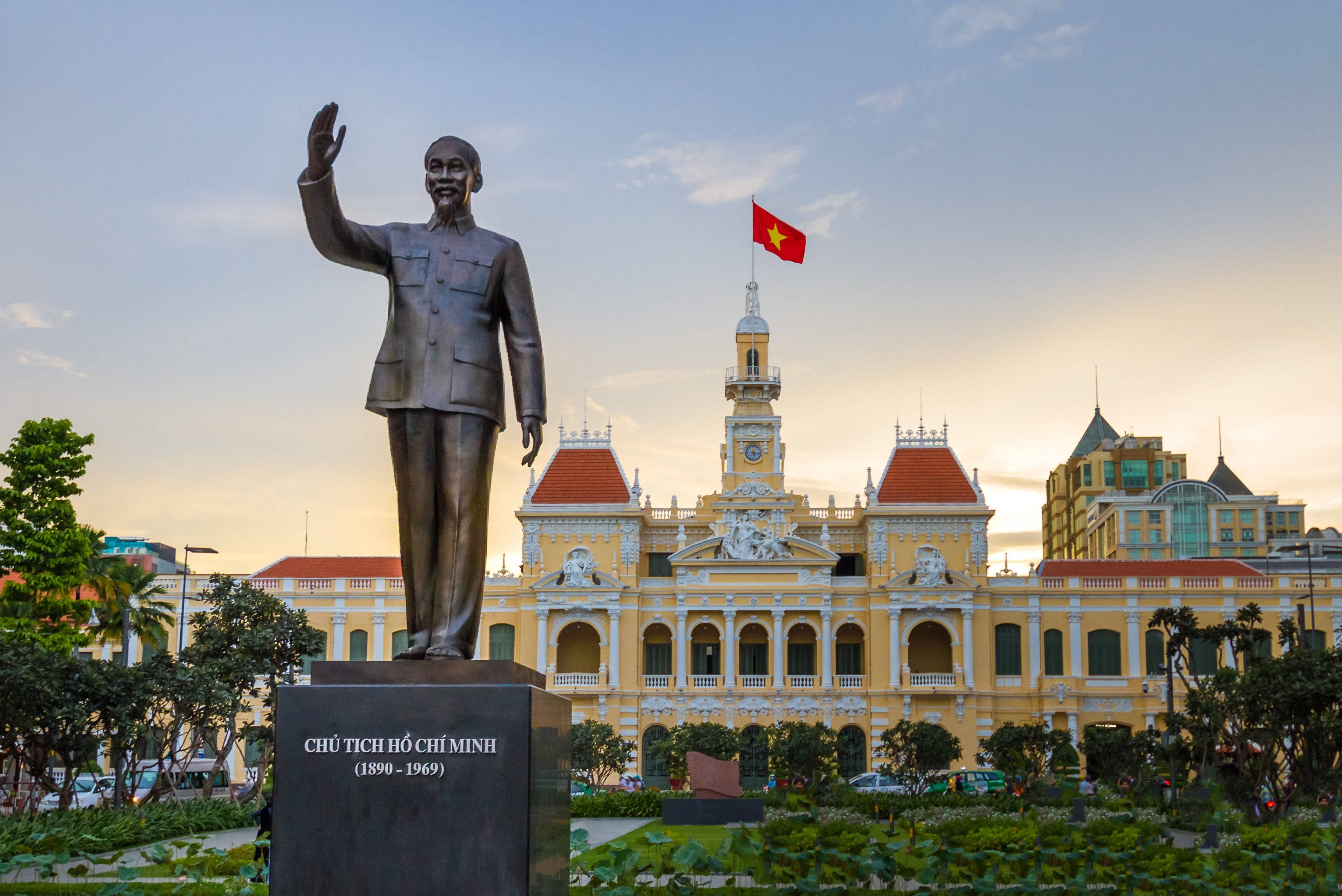 Ho Chi Minh City 桌布 壁紙 — 胡志明銅像在日落時分於一座宏偉的黃色殖民建築旁邊揮手，建築物上飄揚著一面紅旗。