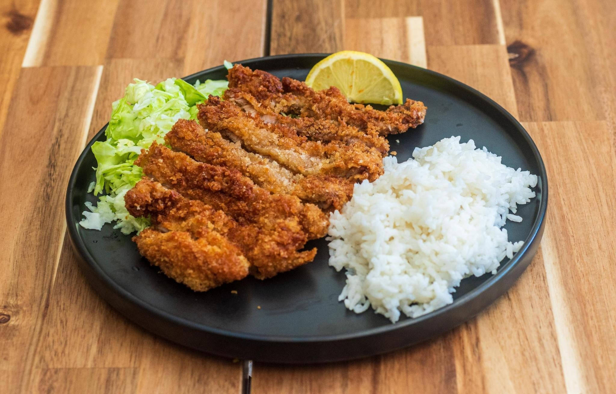 Food Photography 桌布 壁紙 — 脆皮金黃色吉列豬排配白飯和生菜絲，以檸檬角點綴，放在黑色盤子上。