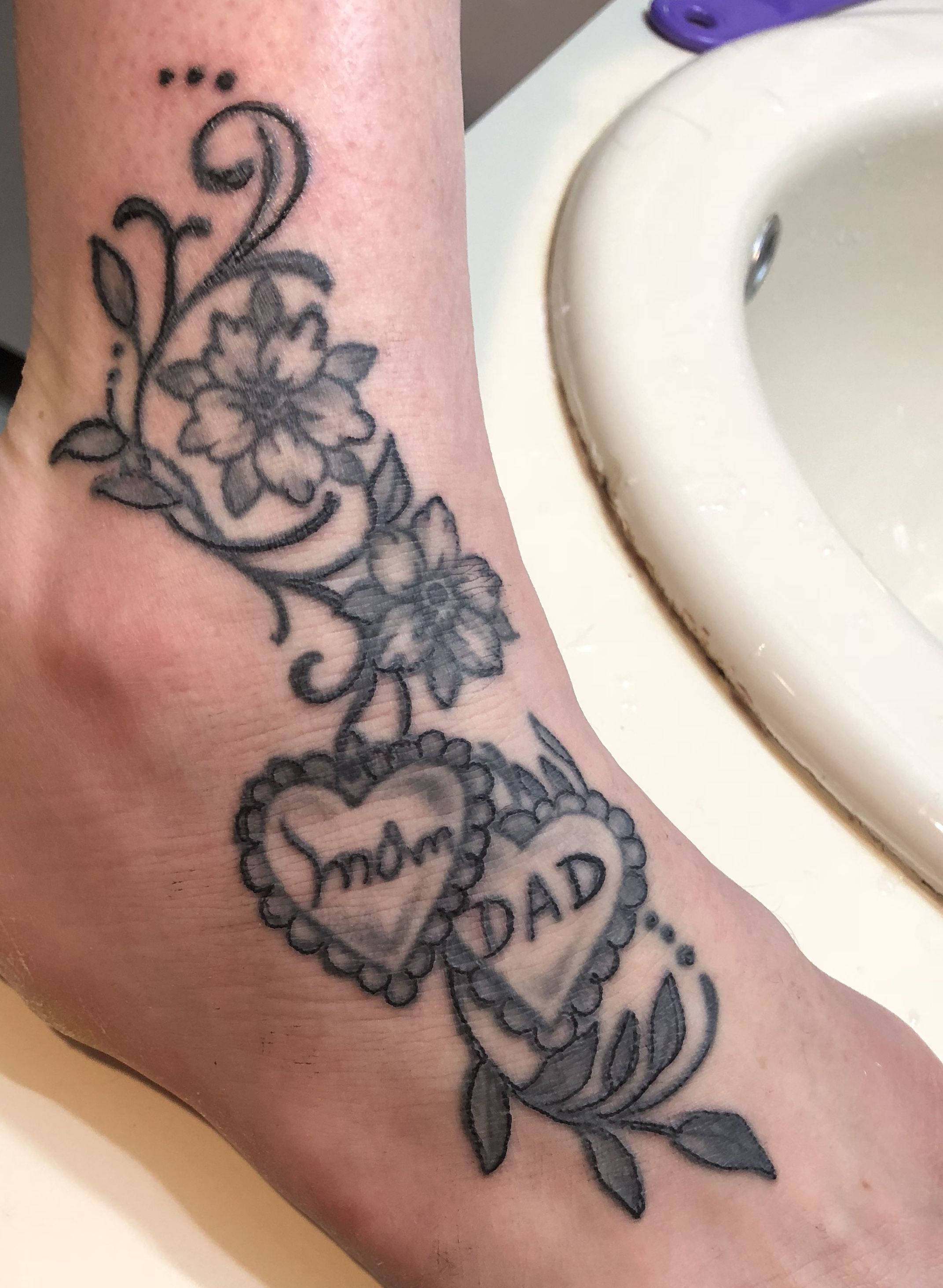 Mom 桌布 壁紙 — 腳踝上的黑灰色紋身，有花卉藤蔓和兩個心形吊飾，上面刻有Mom和Dad。