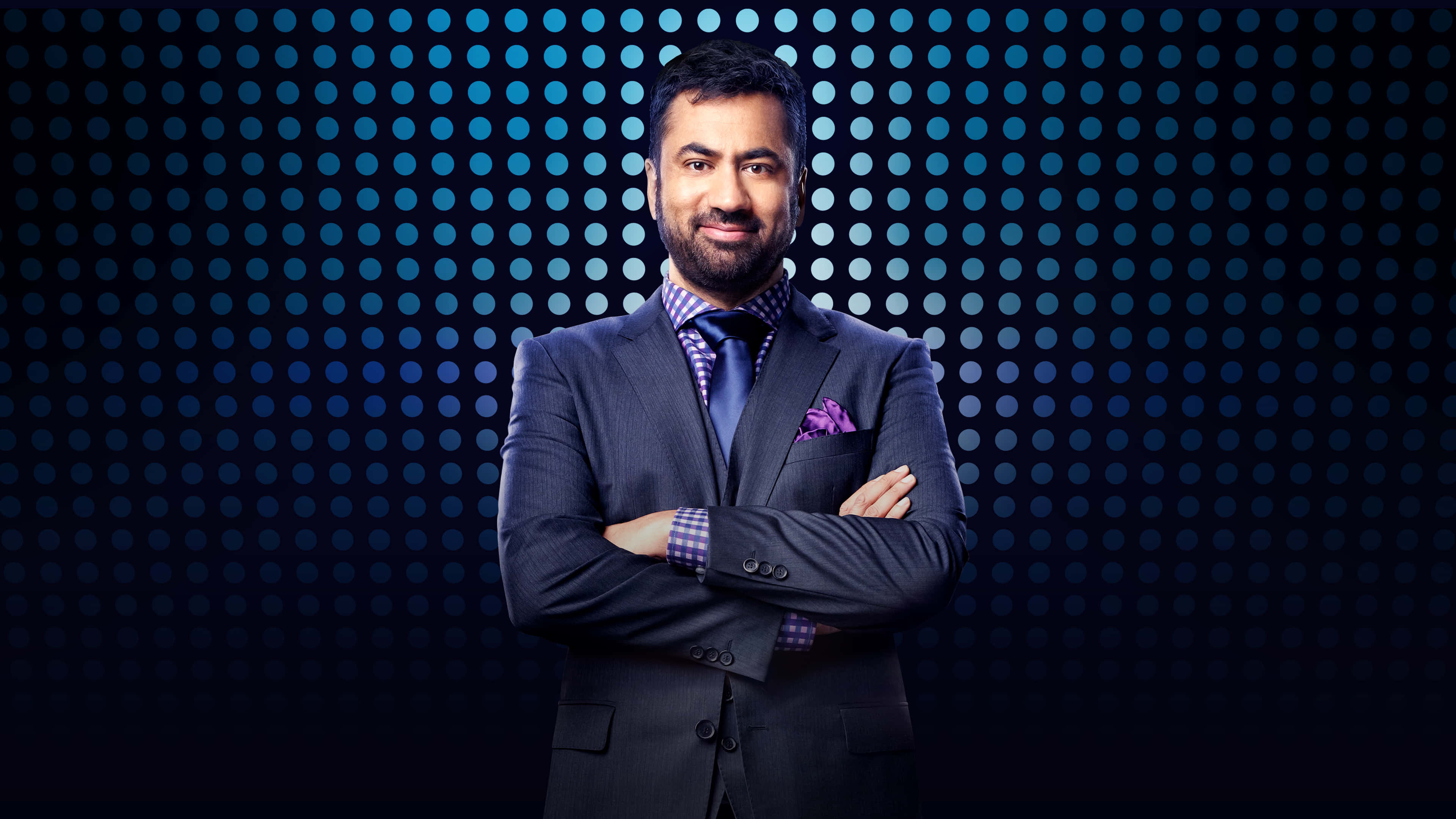 Kal Penn 桌布 壁紙 — 自信滿滿的男士穿著剪裁俐落的西裝，雙手交叉站在發光藍色圓點圖案背景前。