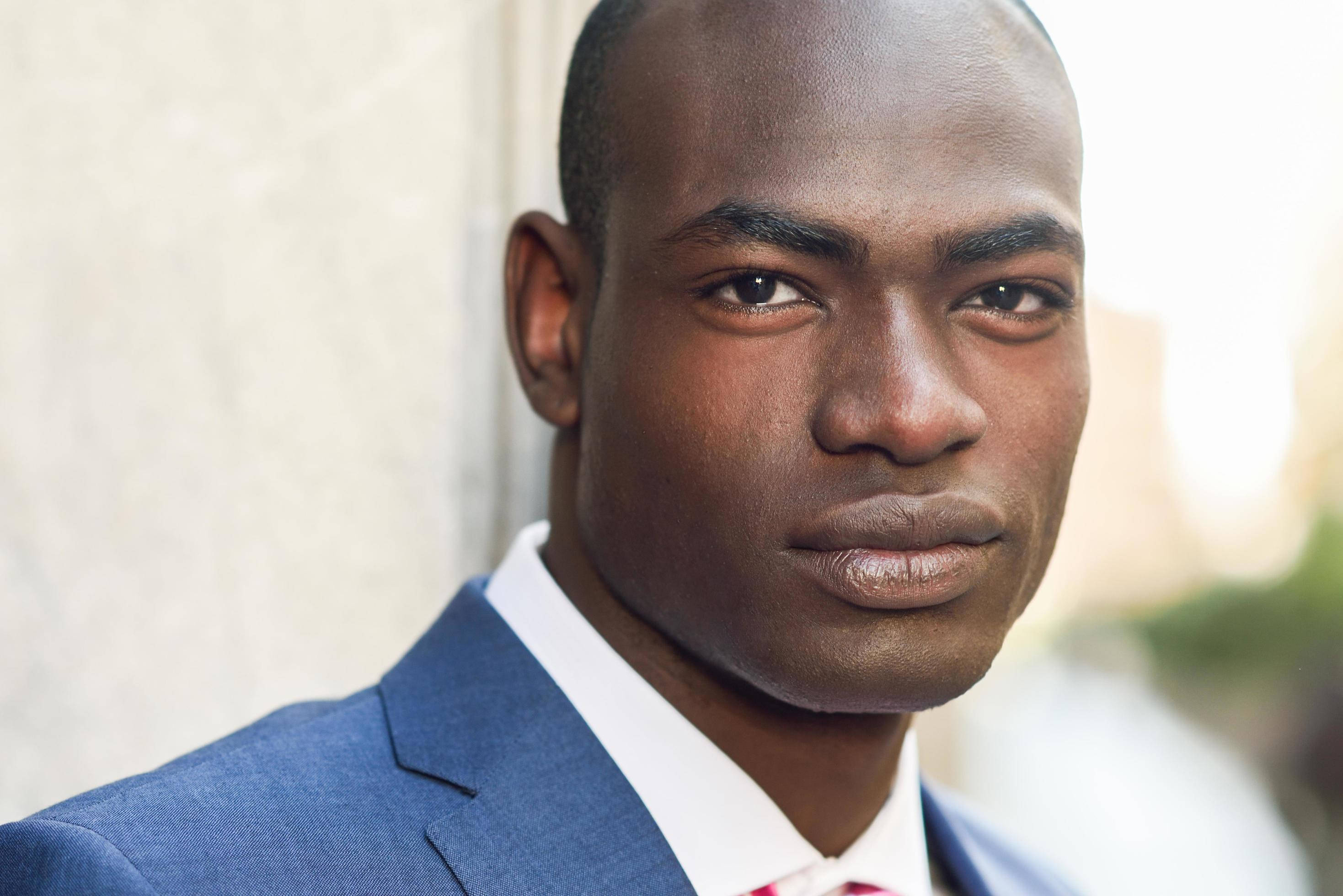 Black Man 桌布 壁紙 — 自信的黑人男子近距離肖像，穿著醒目的藍色西裝和紅色領帶，背景是柔和模糊的城市景觀。