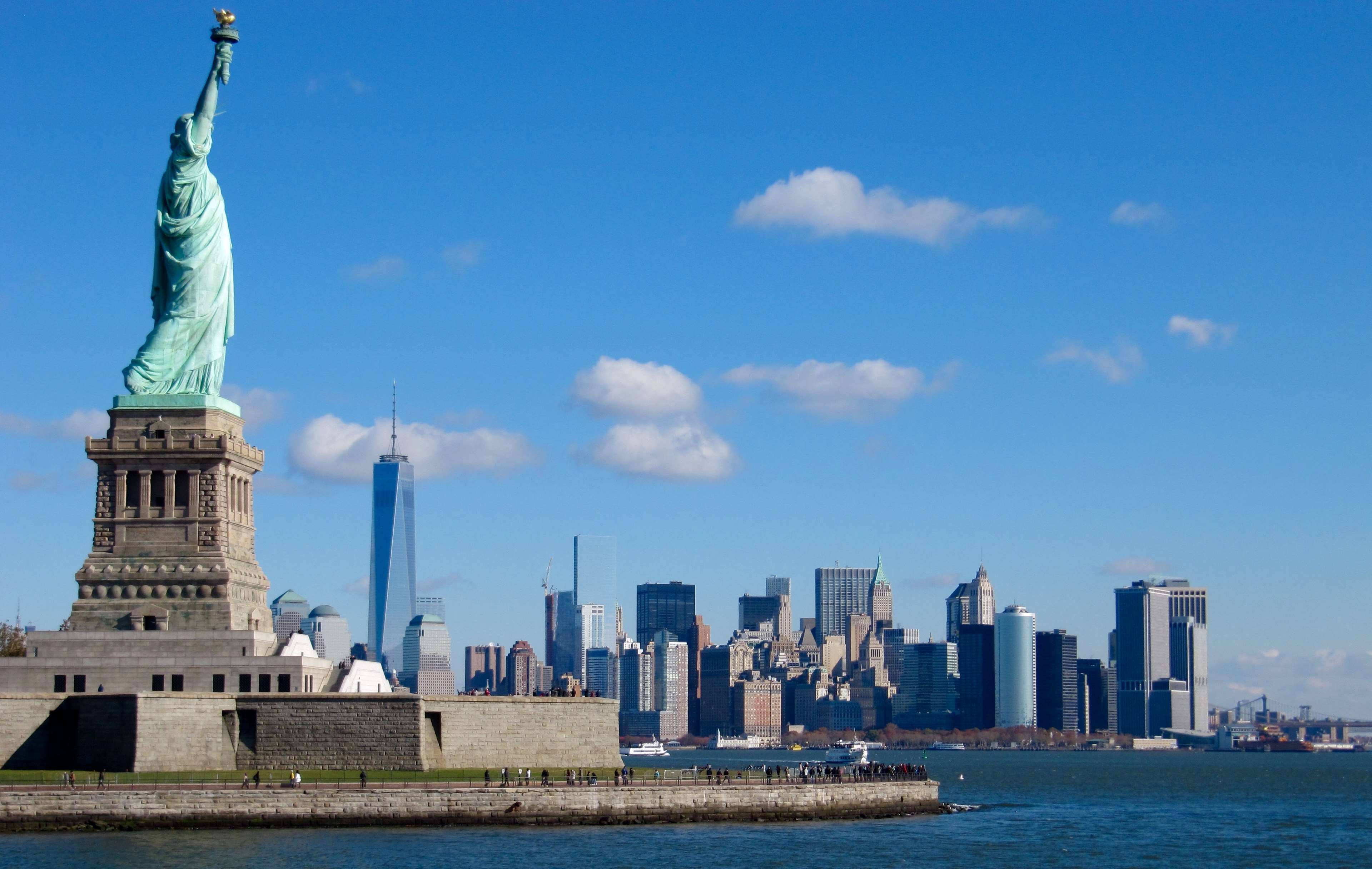 New York 桌布 壁紙 — 自由女神像在晴朗的藍天下高高聳立，背景是現代紐約市天際線和港口。