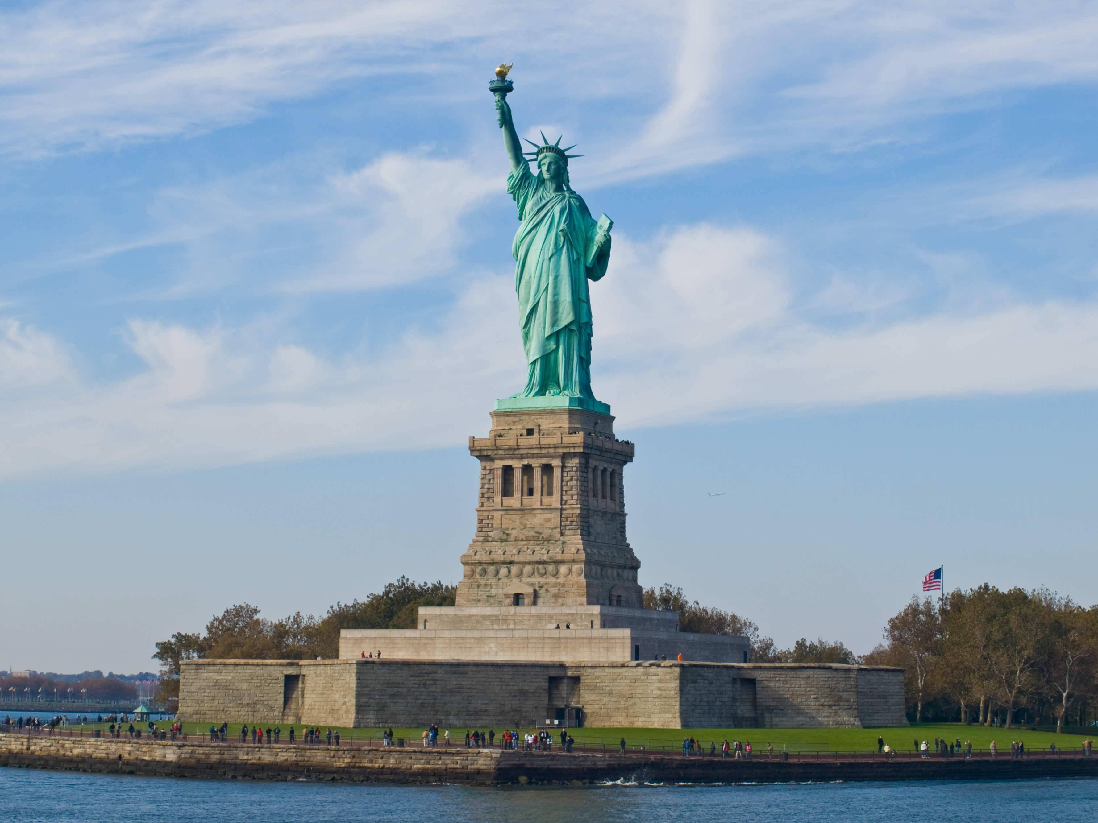 New York 桌布 壁紙 — 自由女神像在自由島上高高聳立，天空湛藍，白雲輕飄，四周環繞著水和遊客。