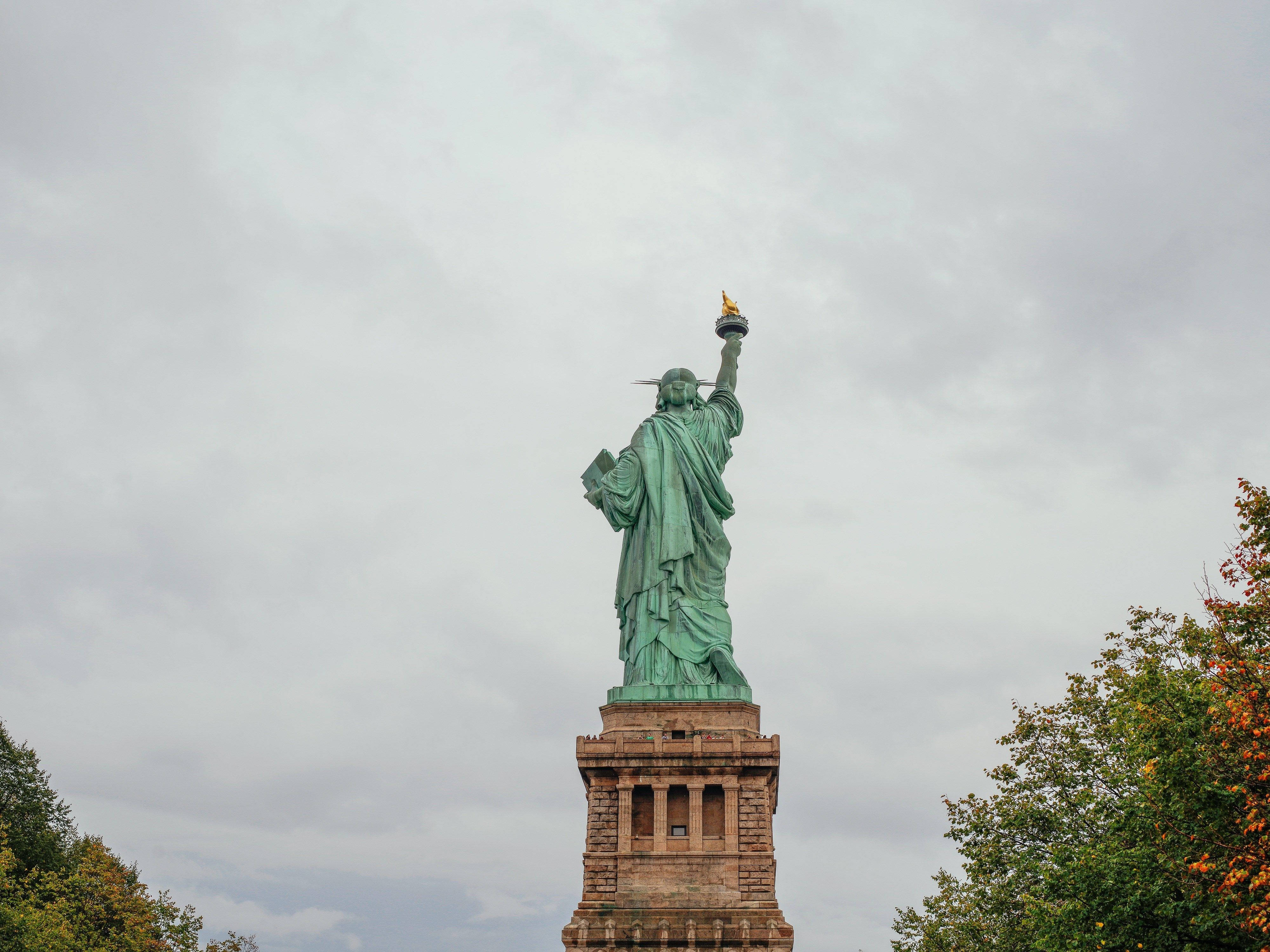 Statue Aesthetic 桌布 壁紙 — 自由女神像在陰雲密布的天空下高高聳立，秋樹環繞著底座。