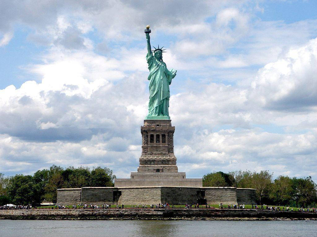 New York 桌布 壁紙 — 自由女神像矗立在基座上，帶有綠色銅鏽，背景是部分多雲的藍天，俯瞰著水域，附近有遊客。