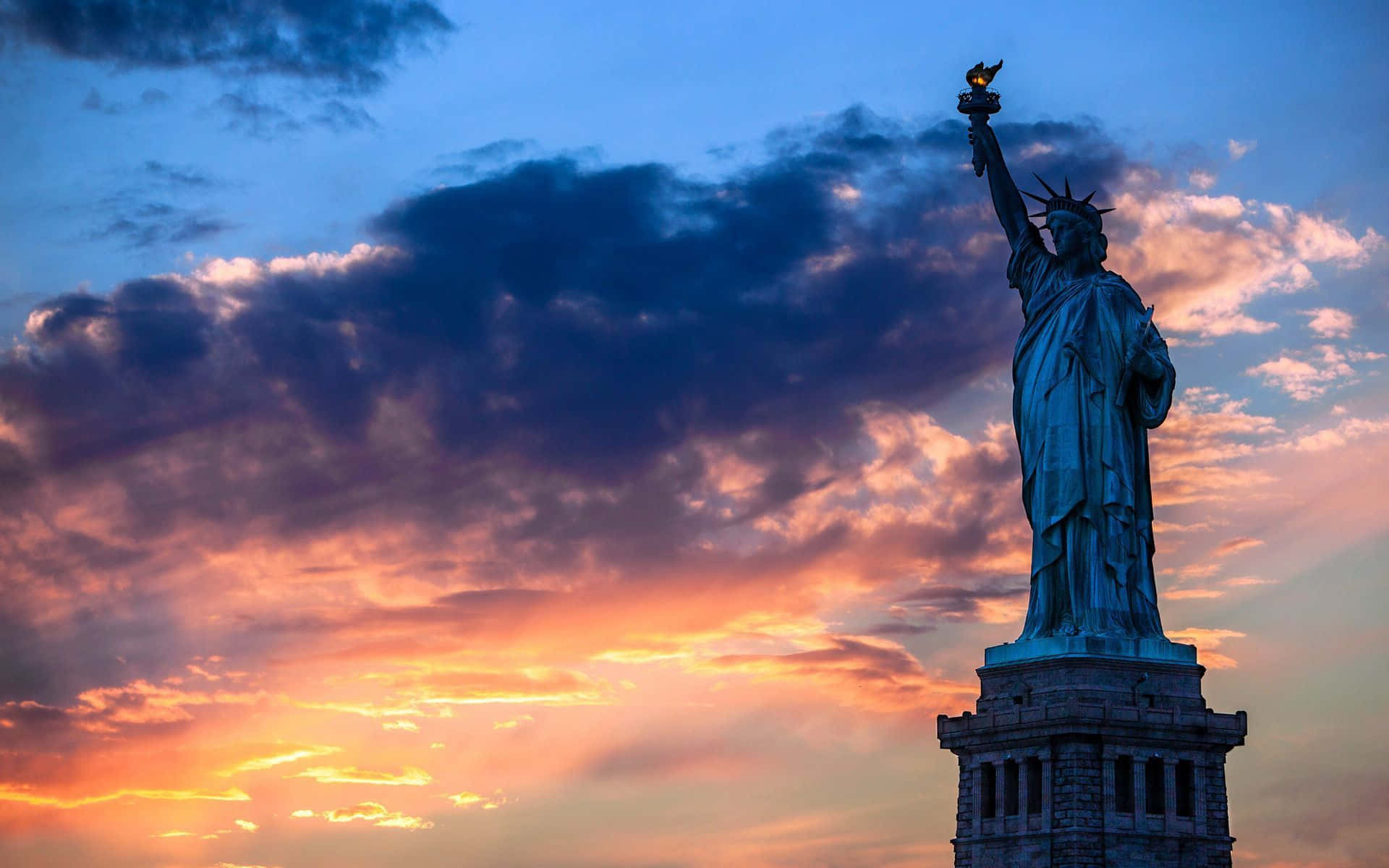 New York 桌布 壁紙 — 自由女神像聳立在戲劇性的暮色天空中，天空被火橙色和深藍色的雲彩塗抹。