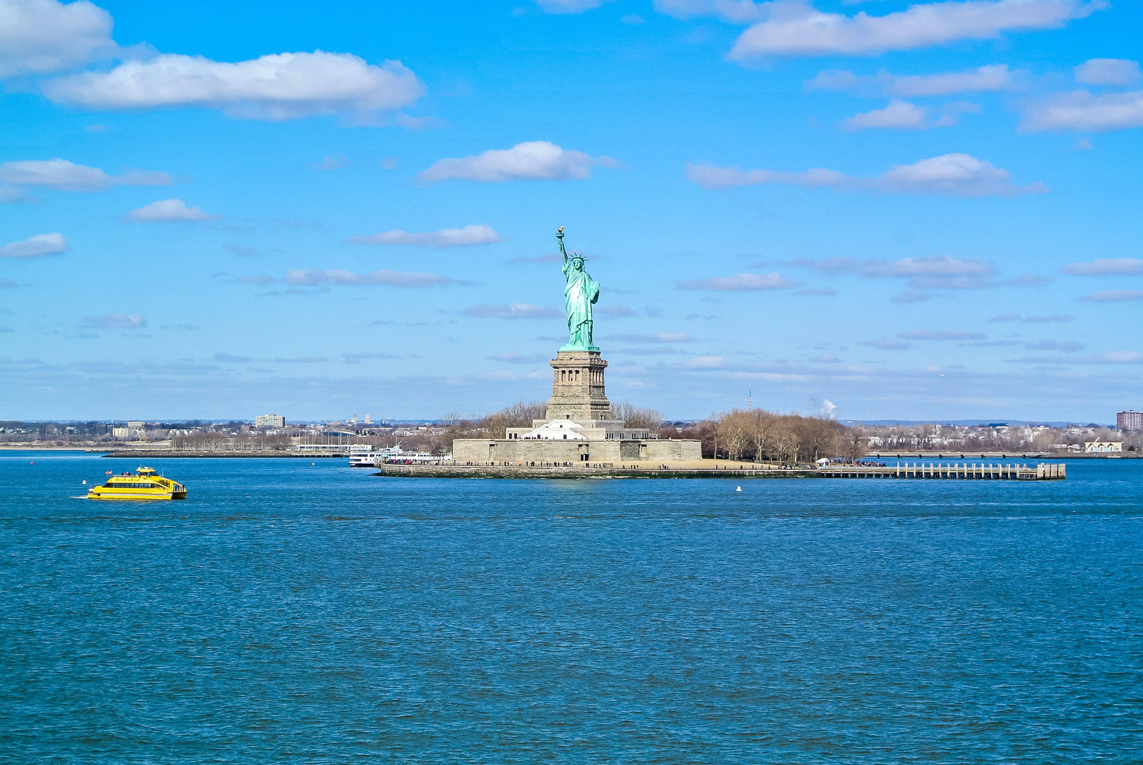 new york 4K 桌布 壁紙 — 自由女神像聳立在自由島上，天空湛藍，白雲點綴，一艘黃色渡輪在深藍色的水面上滑行。