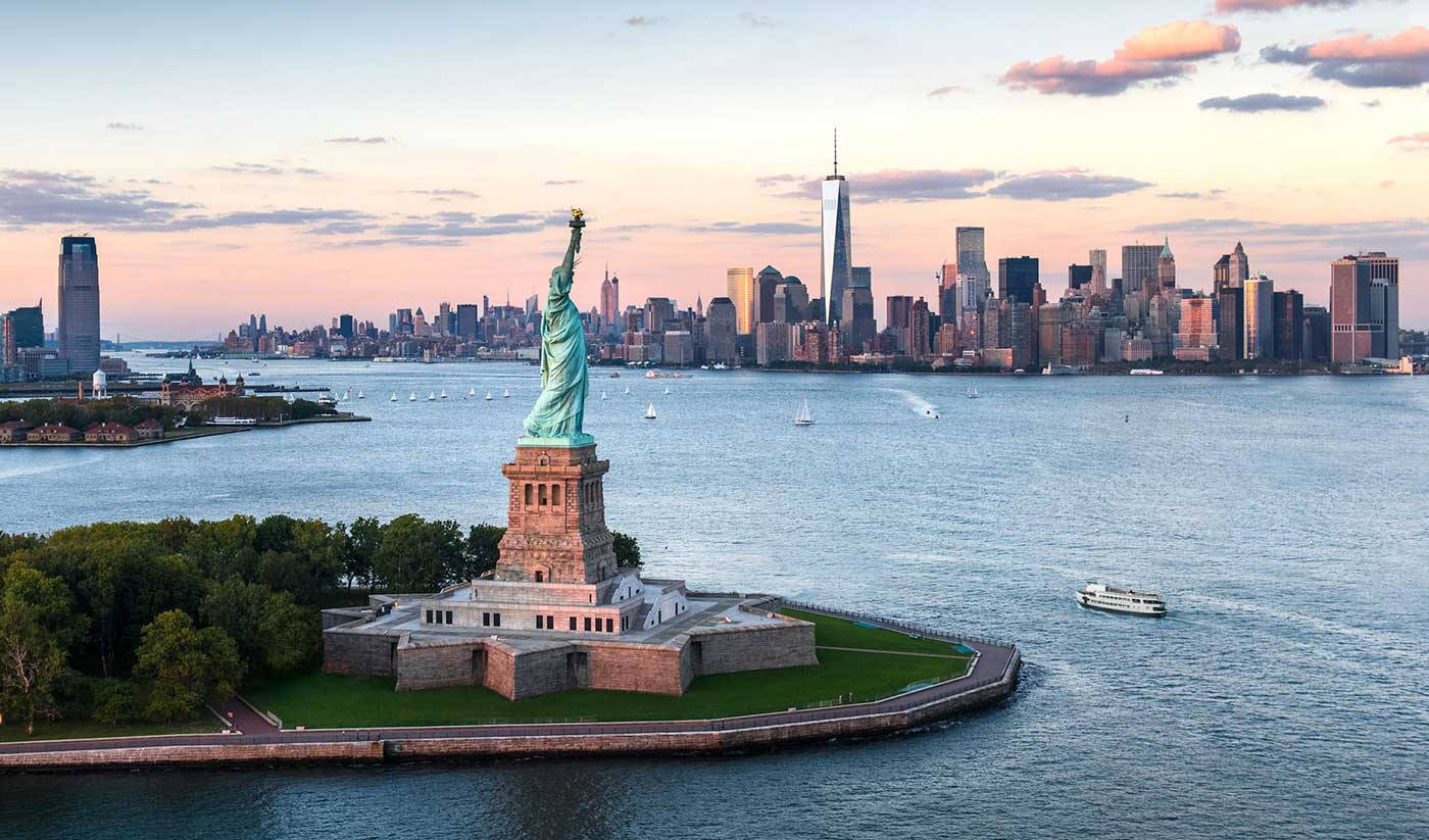 Statue Of Liberty 桌布 壁紙 — 自由女神像聳立在自由島上，紐約市天際線在日落時閃耀光芒。