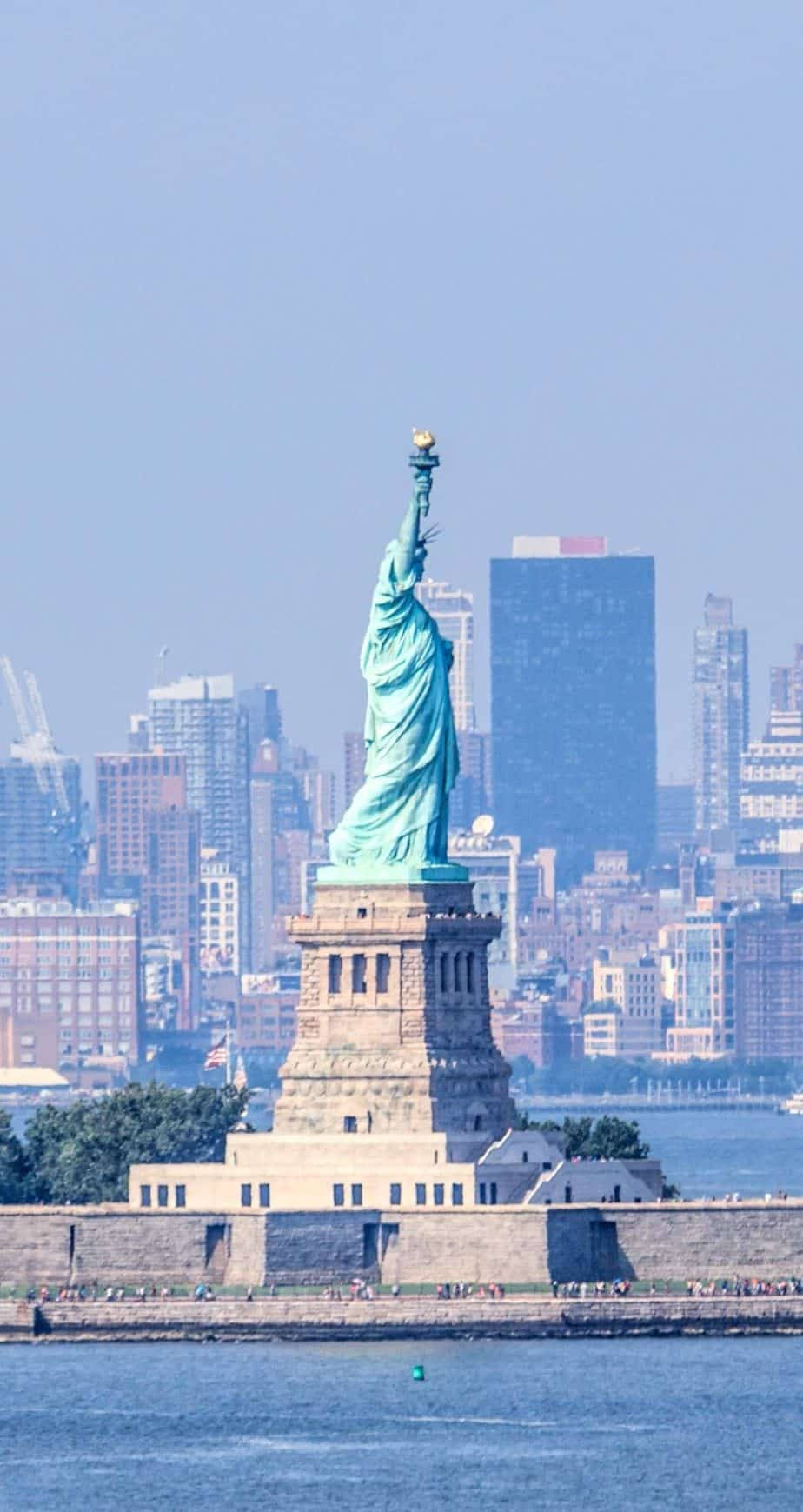 Statue Of Liberty 桌布 壁紙 — 自由女神像聳立在自由島上，背景是朦朧的紐約市天際線，上方是清澈的藍天。