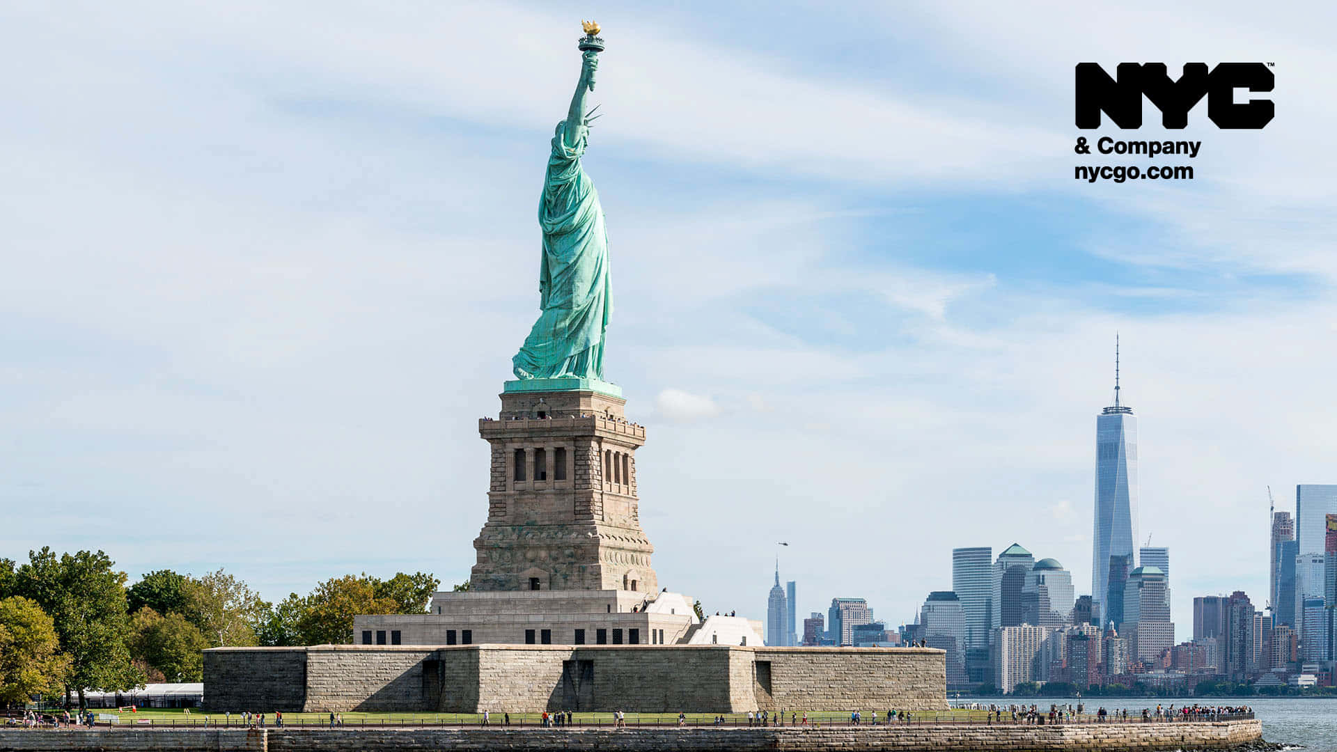 Statue Of Liberty 桌布 壁紙 — 自由女神像高聳入雲，火炬高舉在藍天下，俯瞰紐約市天際線，可見世界貿易中心一號大樓。