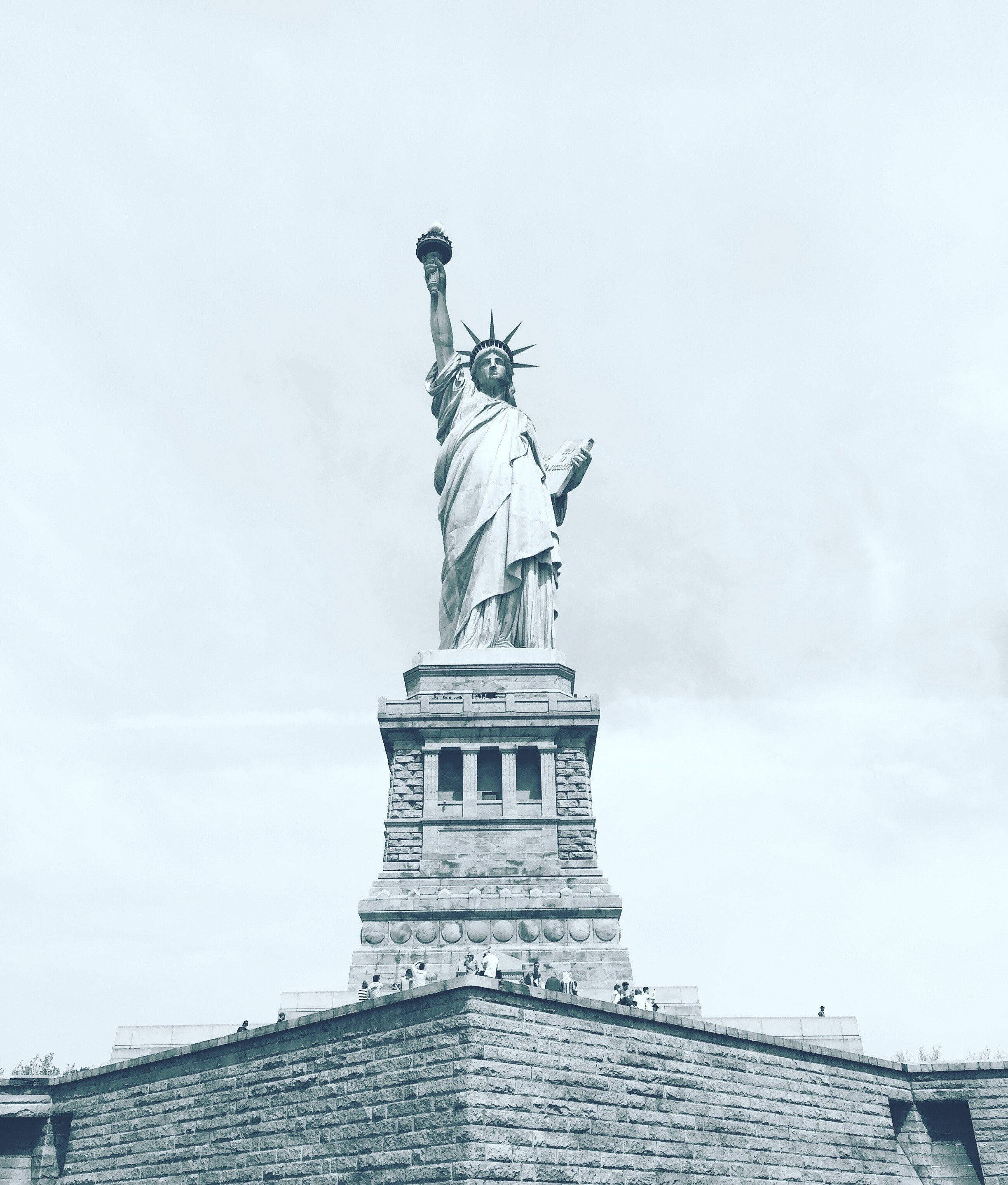 Statue Aesthetic 桌布 壁紙 — 自由女神像高舉火炬，背景為多雲天空，基座上有遊客的黑白影像。