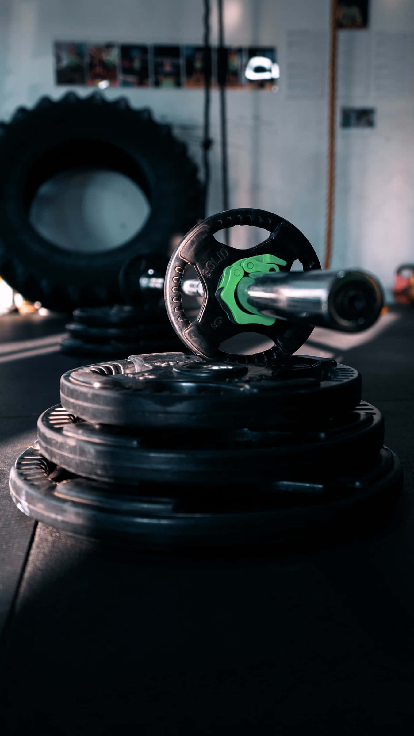 Barbell 桌布 壁紙 — 舉重槓鈴的特寫，綠色槓鈴片放在一堆黑色槓鈴片上，位於燈光昏暗的健身房內。