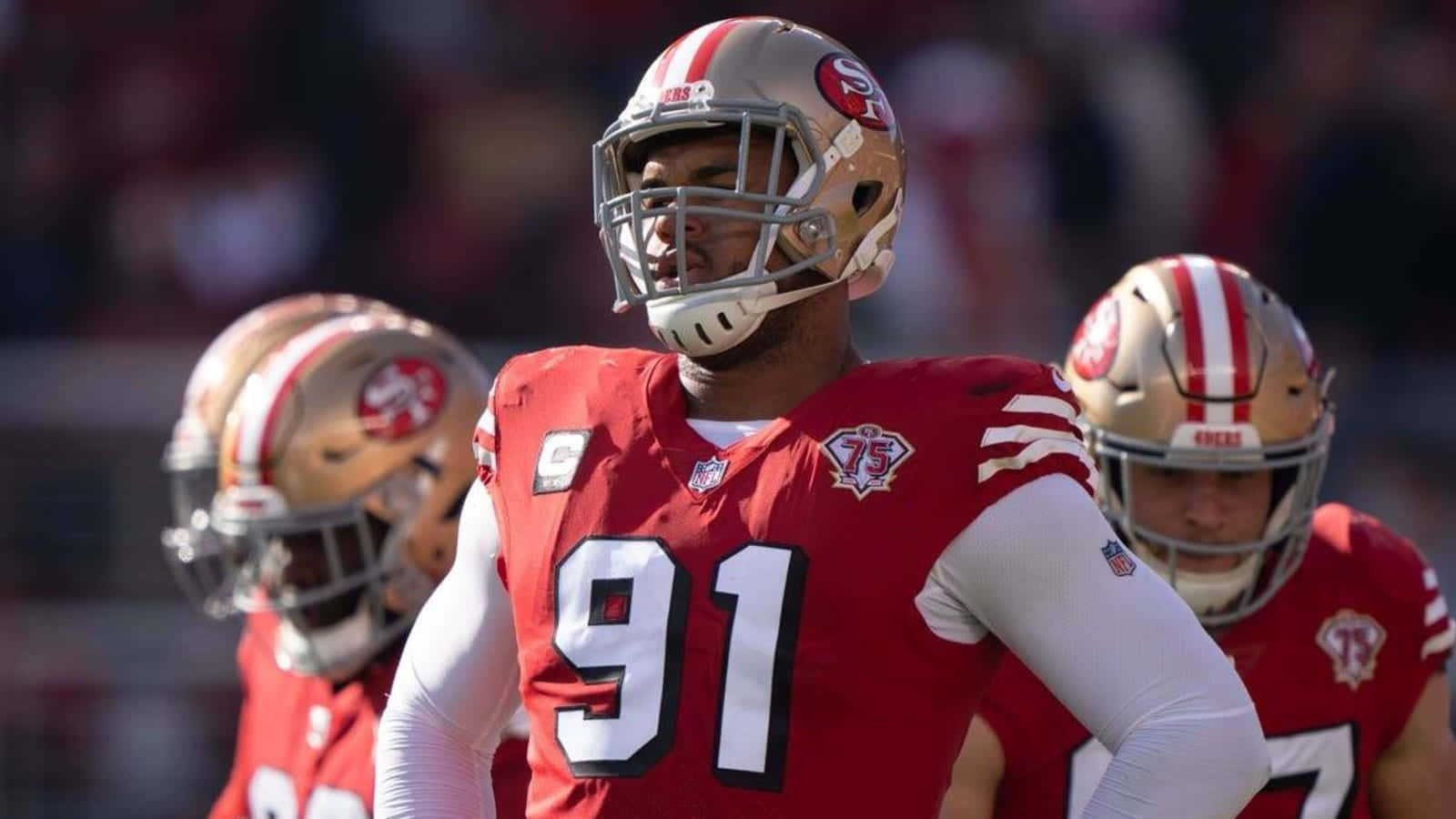 San Francisco 49ers 桌布 壁紙 — 舊金山49人美式足球員身穿紅色和金色球衣，在陽光明媚的比賽中站在球場上。