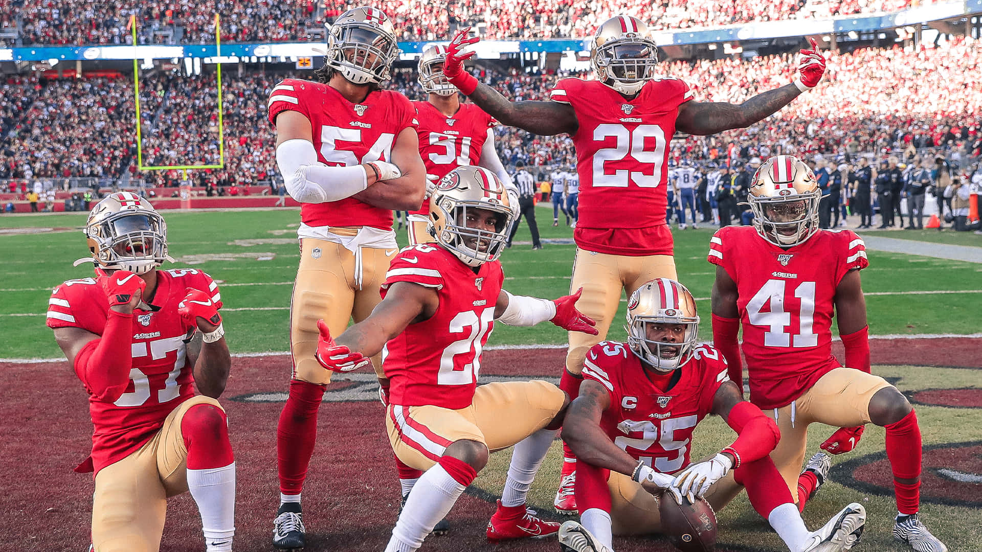 49ers 桌布 壁紙 — 舊金山49人隊美式足球員身穿紅色和金色球衣，在陽光明媚的比賽日於擠滿觀眾的球場上自信地擺姿勢。
