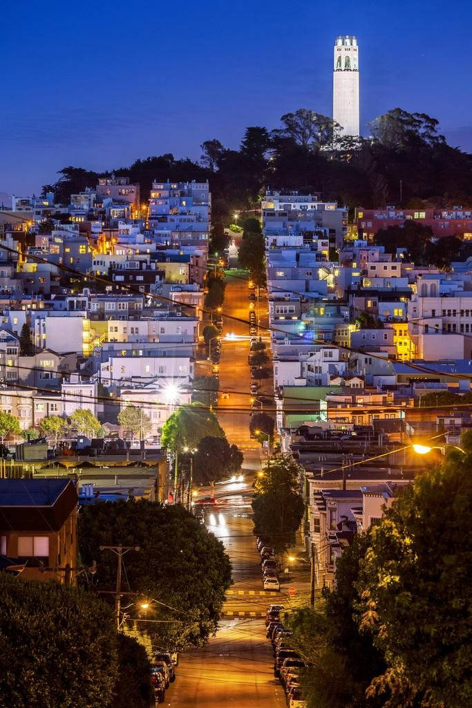 san francisco iPhone 桌布 壁紙 — 舊金山一條陡峭的街道，在暮色中閃爍著街燈，科伊特塔聳立在山邊房屋之上。