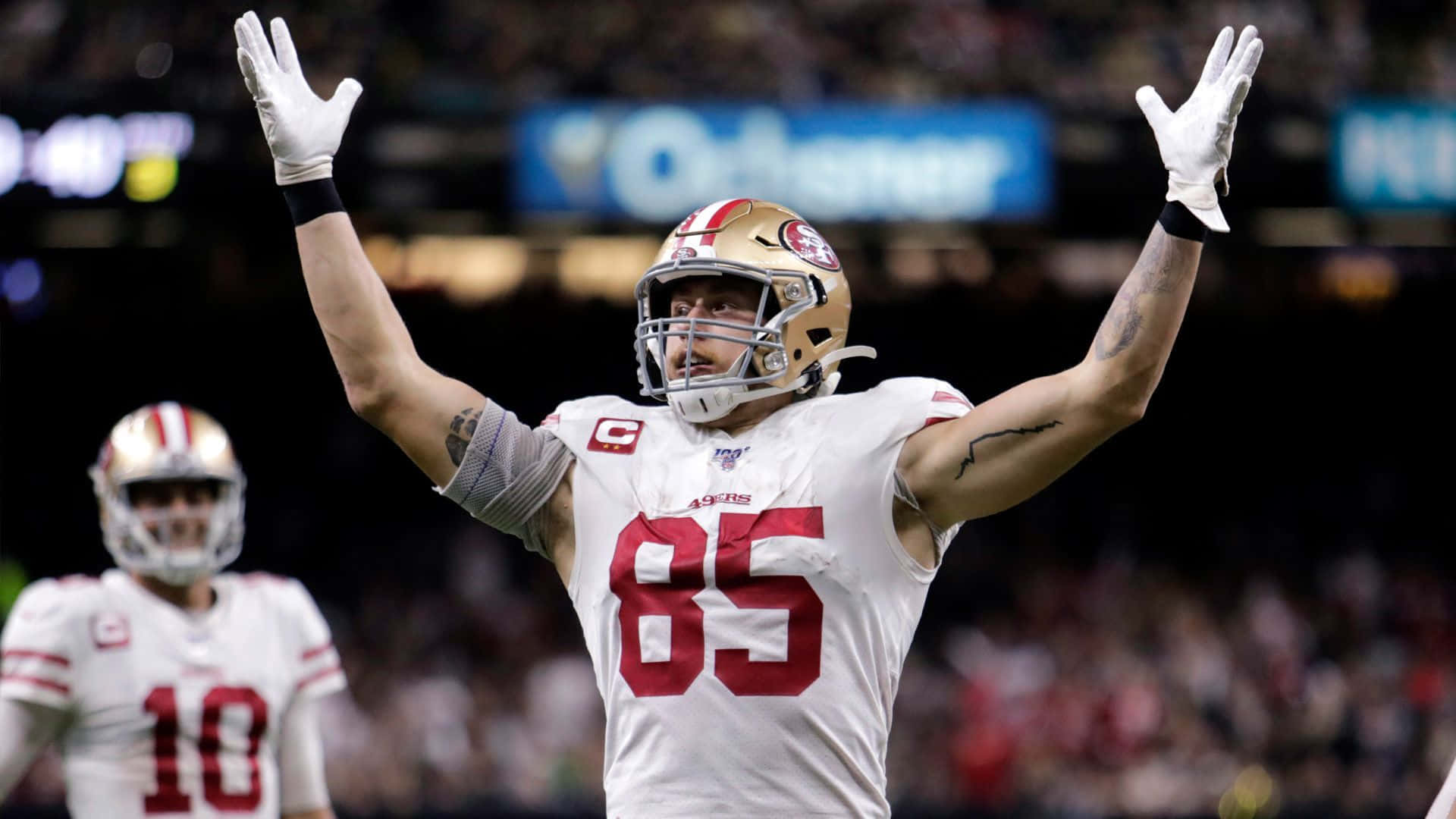 George Kittle 桌布 壁紙 — 舊金山四九人美式足球員，身穿白色球衣85號，在球場燈光下高舉雙臂慶祝勝利。