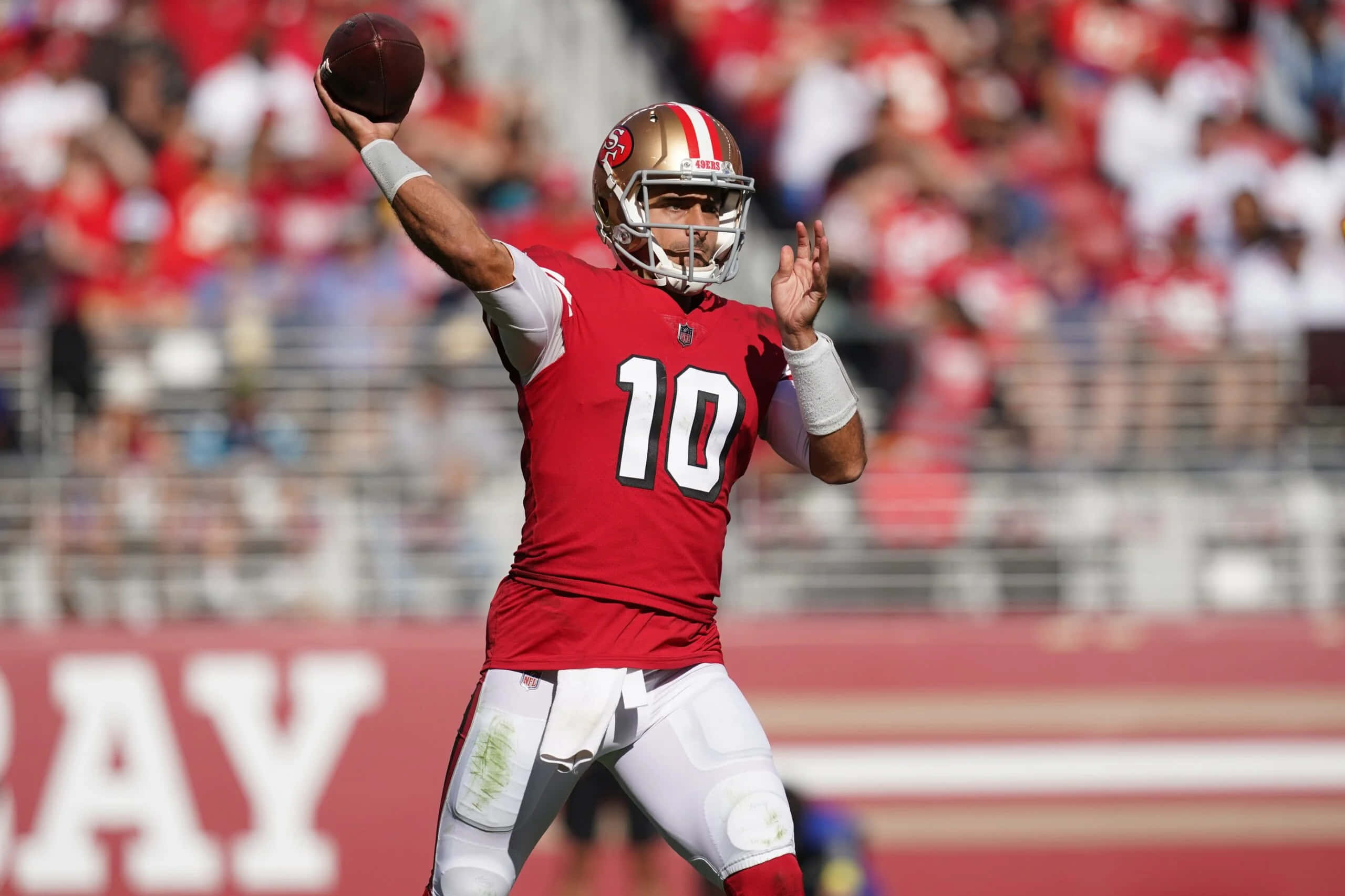 Jimmy Garoppolo 桌布 壁紙 — 舊金山四九人隊的四分衛身穿紅色10號球衣，在日間比賽中擲出橄欖球，背景是模糊的球場觀眾。