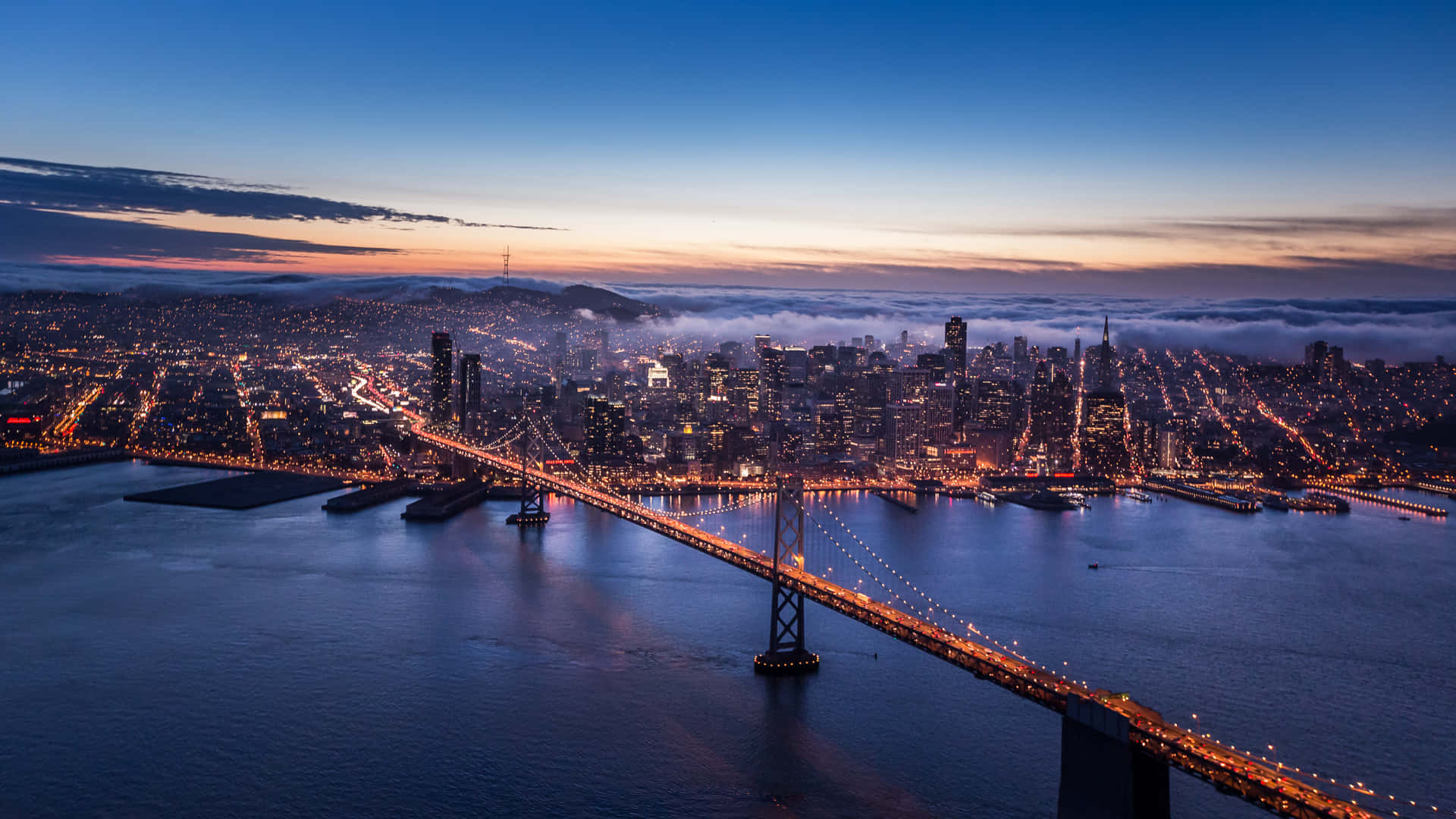 San Francisco 桌布 壁紙 — 舊金山黃昏時的鳥瞰圖，海灣大橋在水面上閃耀，城市燈光穿透霧氣。
