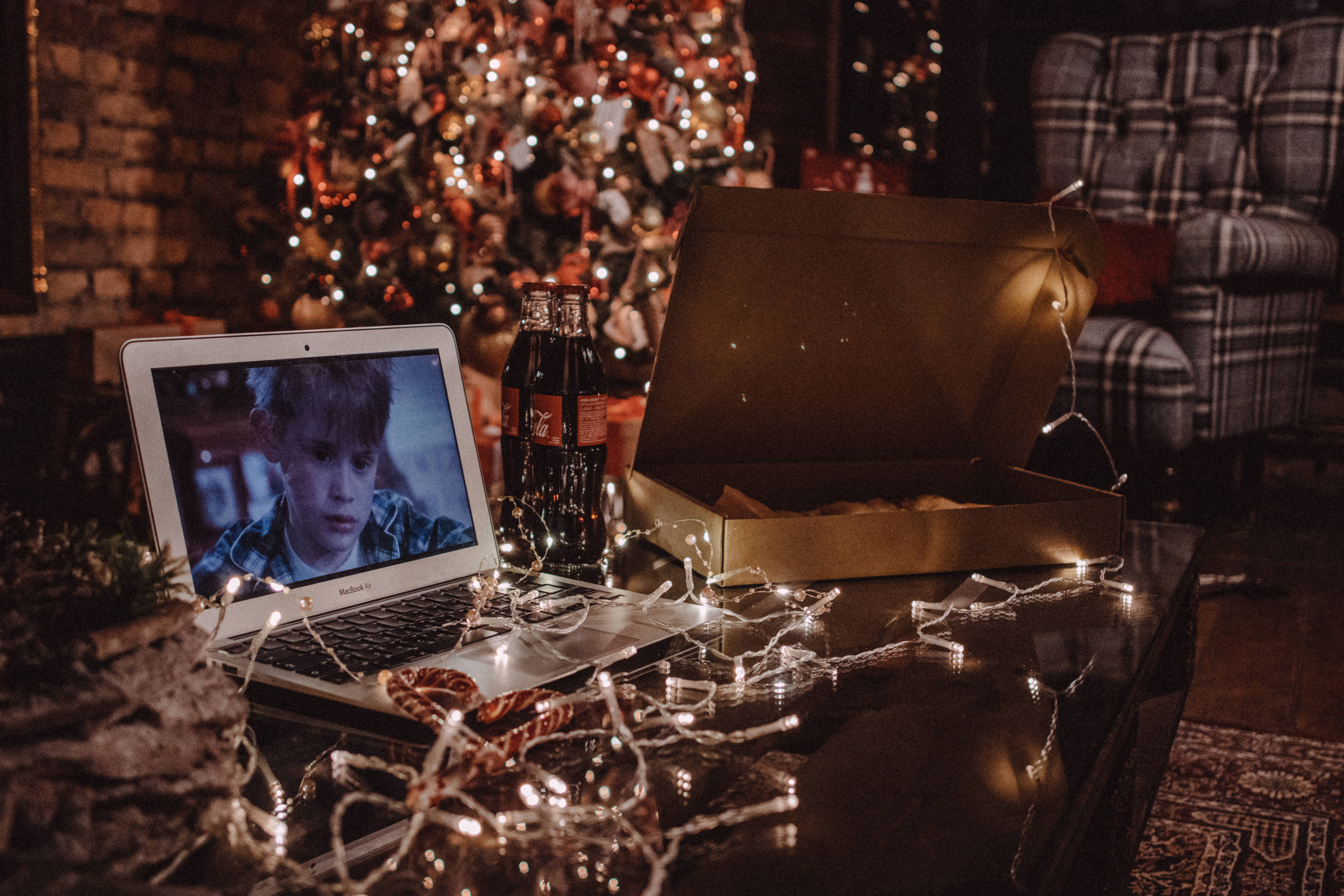 christmas laptop 桌布 壁紙 — 舒適的假日夜晚場景，咖啡桌上放著顯示電影的筆記型電腦、可口可樂、披薩盒和閃爍的仙女燈。