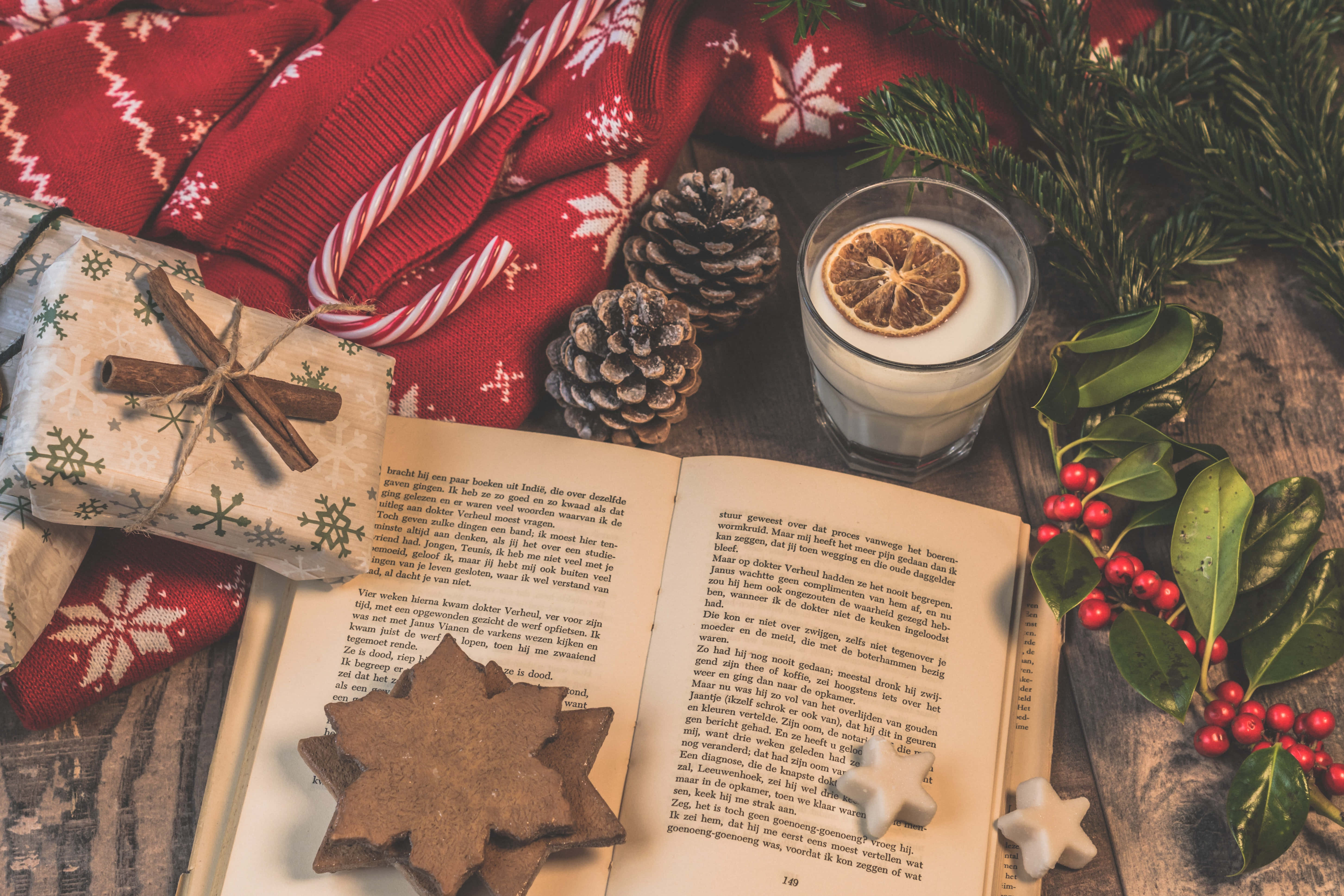 Christmas Scene 桌布 壁紙 — 舒適的冬季假期場景，有打開的書本、松果、肉桂棒和節日紅色毛衣，放在質樸的木桌上。